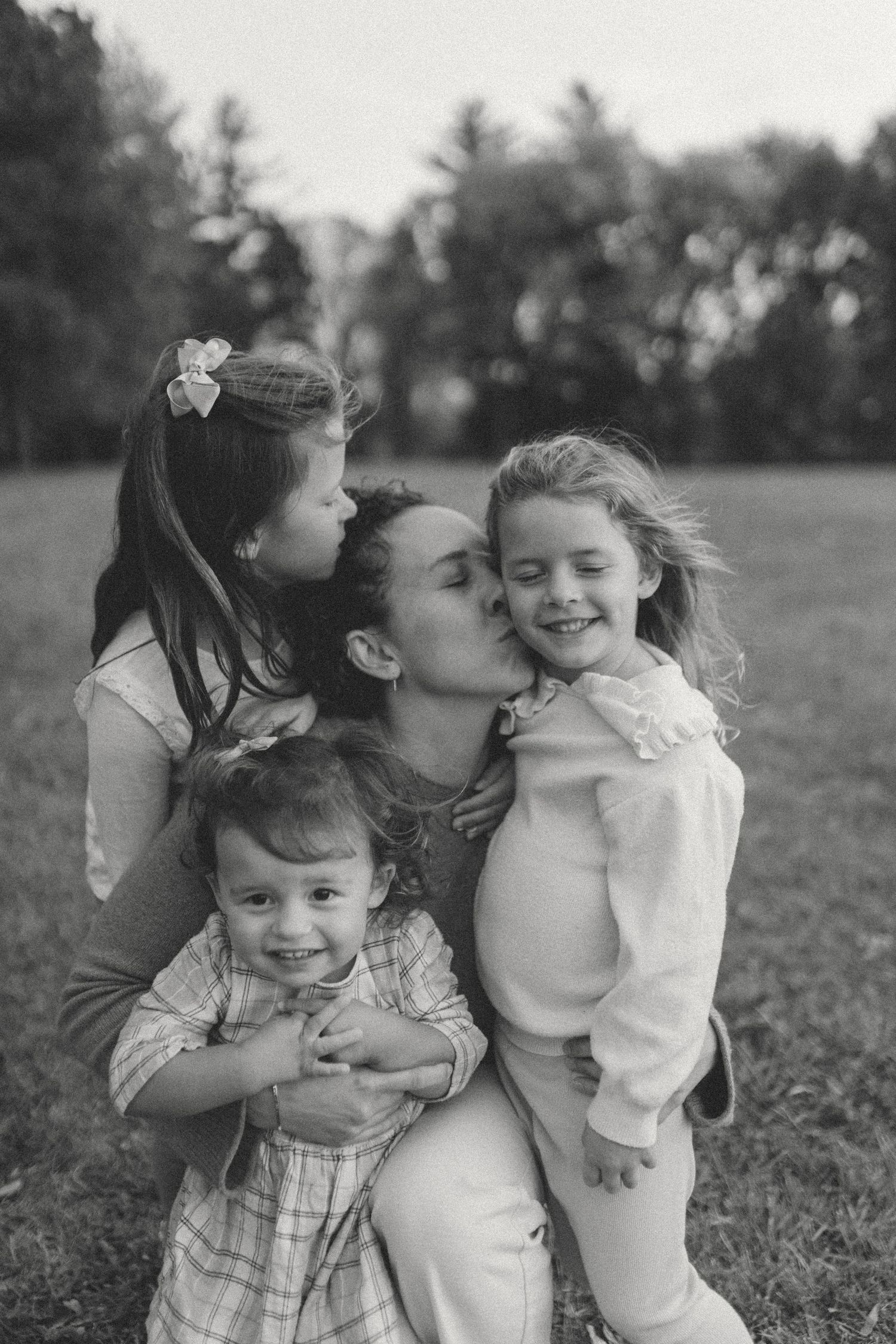 Smiling candid photo, documentary style, of a mother with three daughters laughing and cuddling in a field