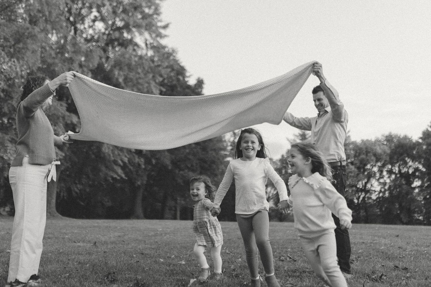 Family playing in a field with three kids running under a blanket while parents smile and watch them enjoy the fun.