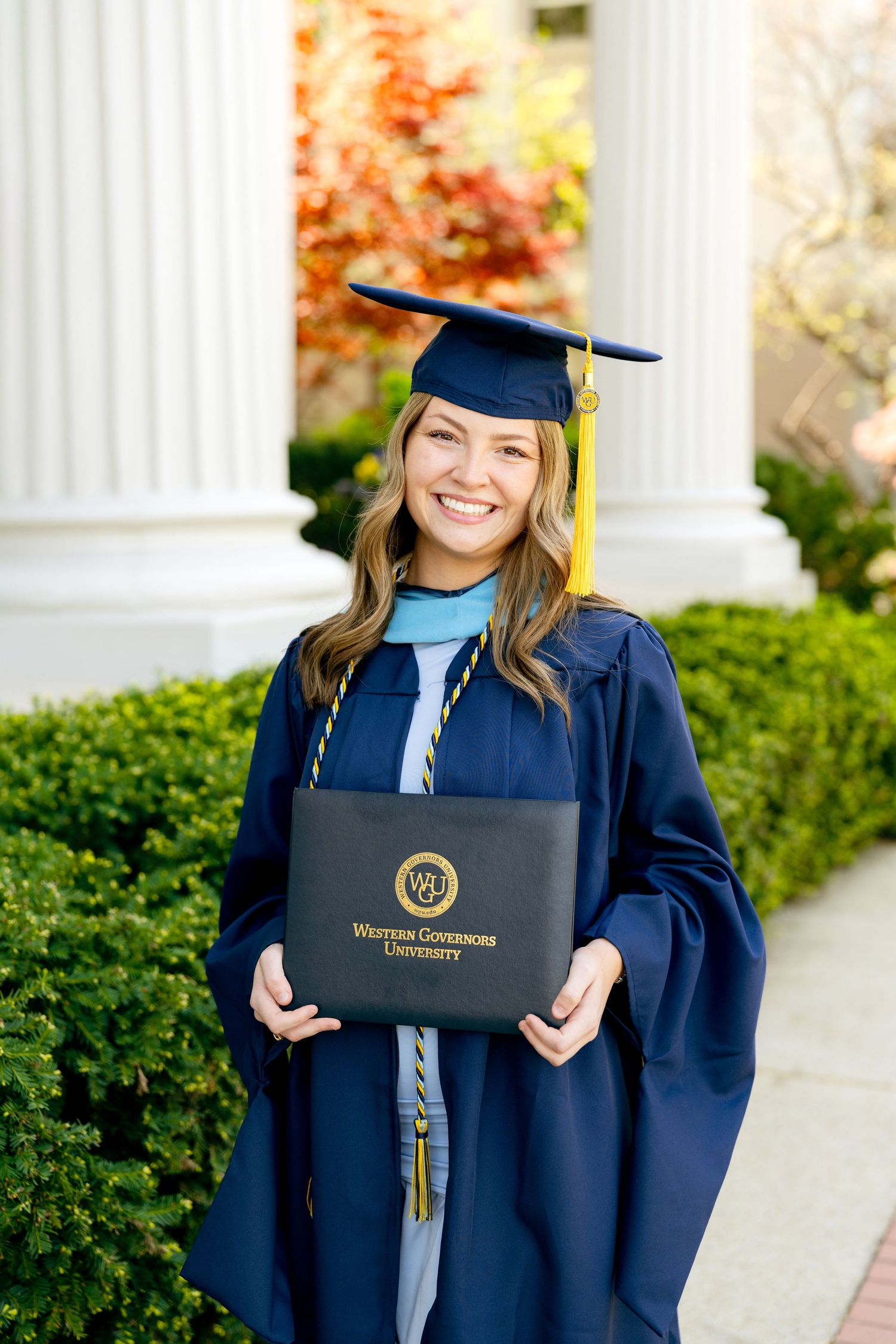 Graduate in blue regalia displaying diploma holder in front of white columns at Belmont University.