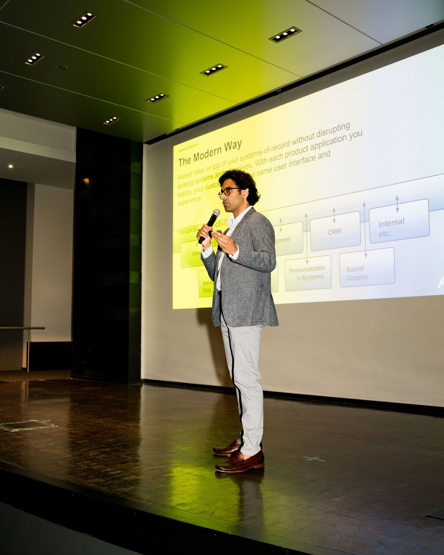 A presenter in a gray blazer and light pants speaks at a podium during a business presentation with projected slides.