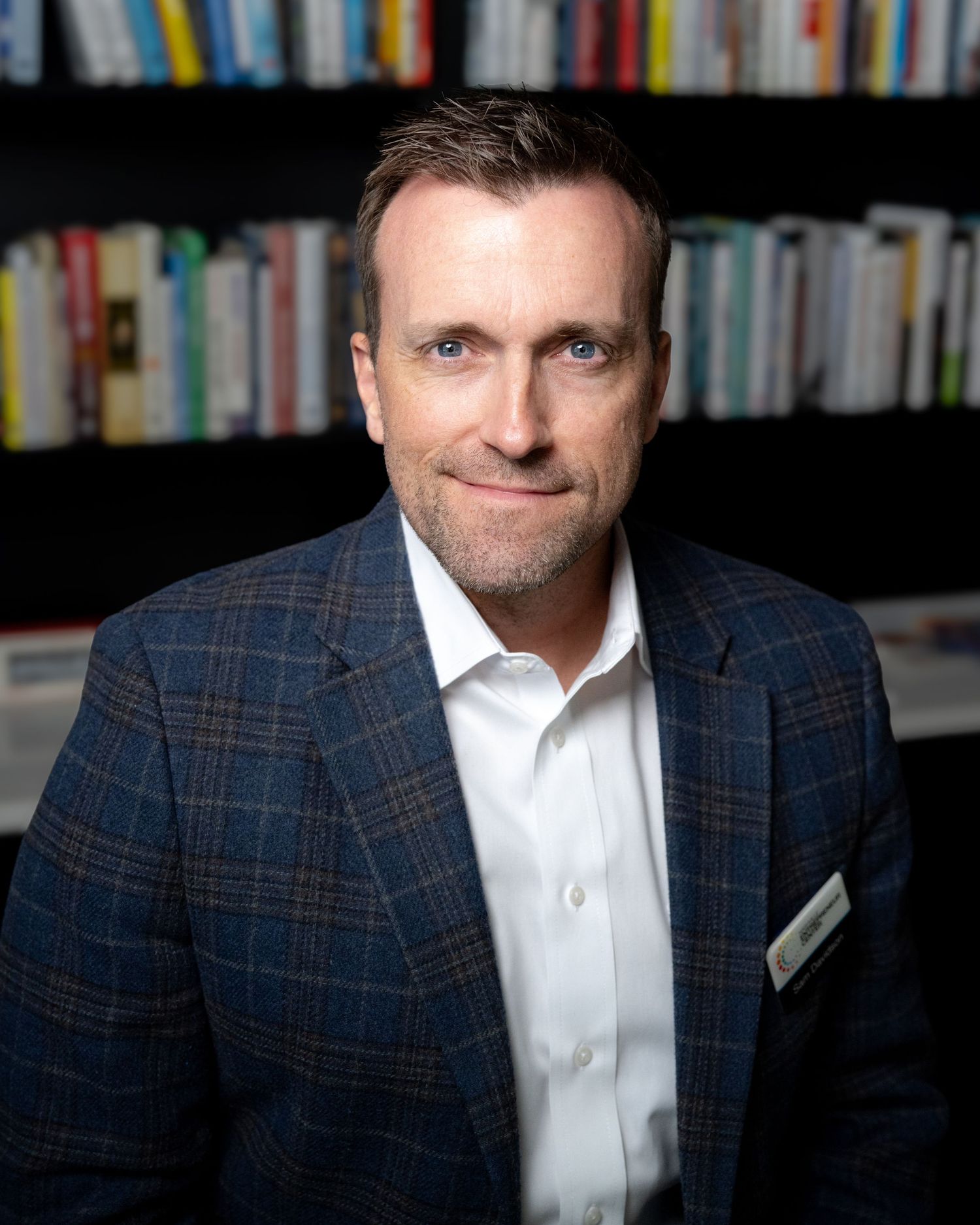A smiling person in a navy plaid blazer and white shirt sits in front of a bookshelf in a library setting.