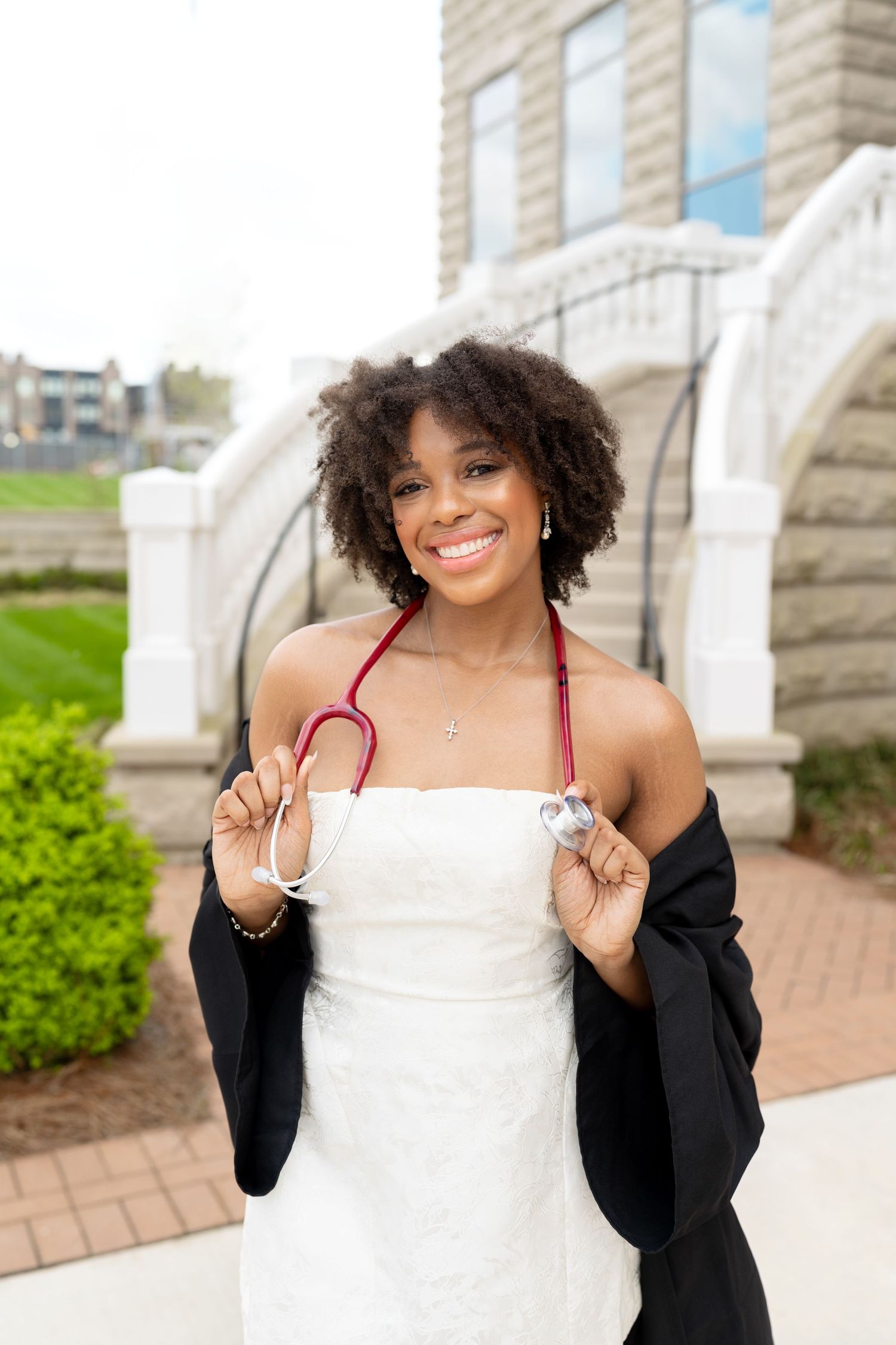 Graduate celebrates in white dress and black graduation gown against stone building backdrop.