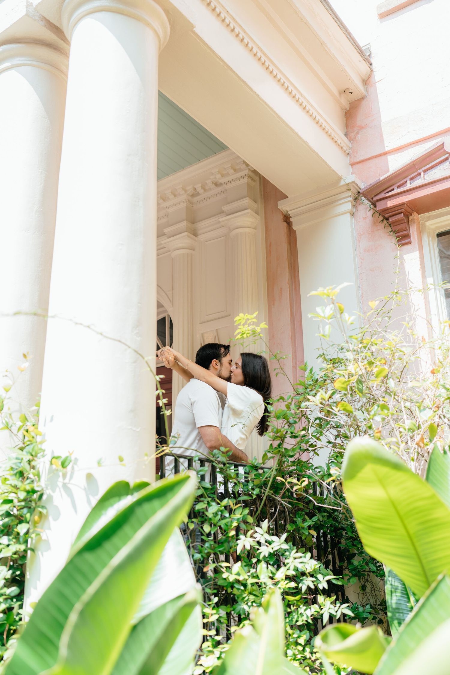 A couple embraces under elegant white columns surrounded by tropical plants.