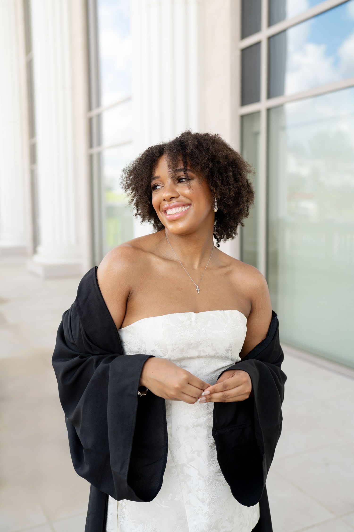A Belmont Grad in a white dress and black gown poses playfully against a bright building exterior in downtown nashville.