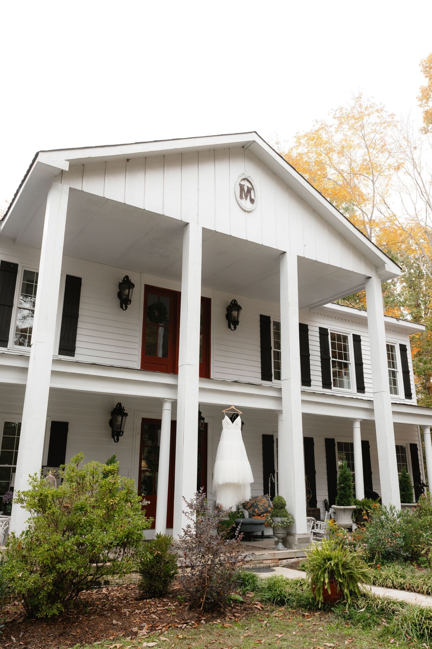 White colonial wedding venue with large pillars, black shutters, and a bridal gown hanging from the porch in autumn.