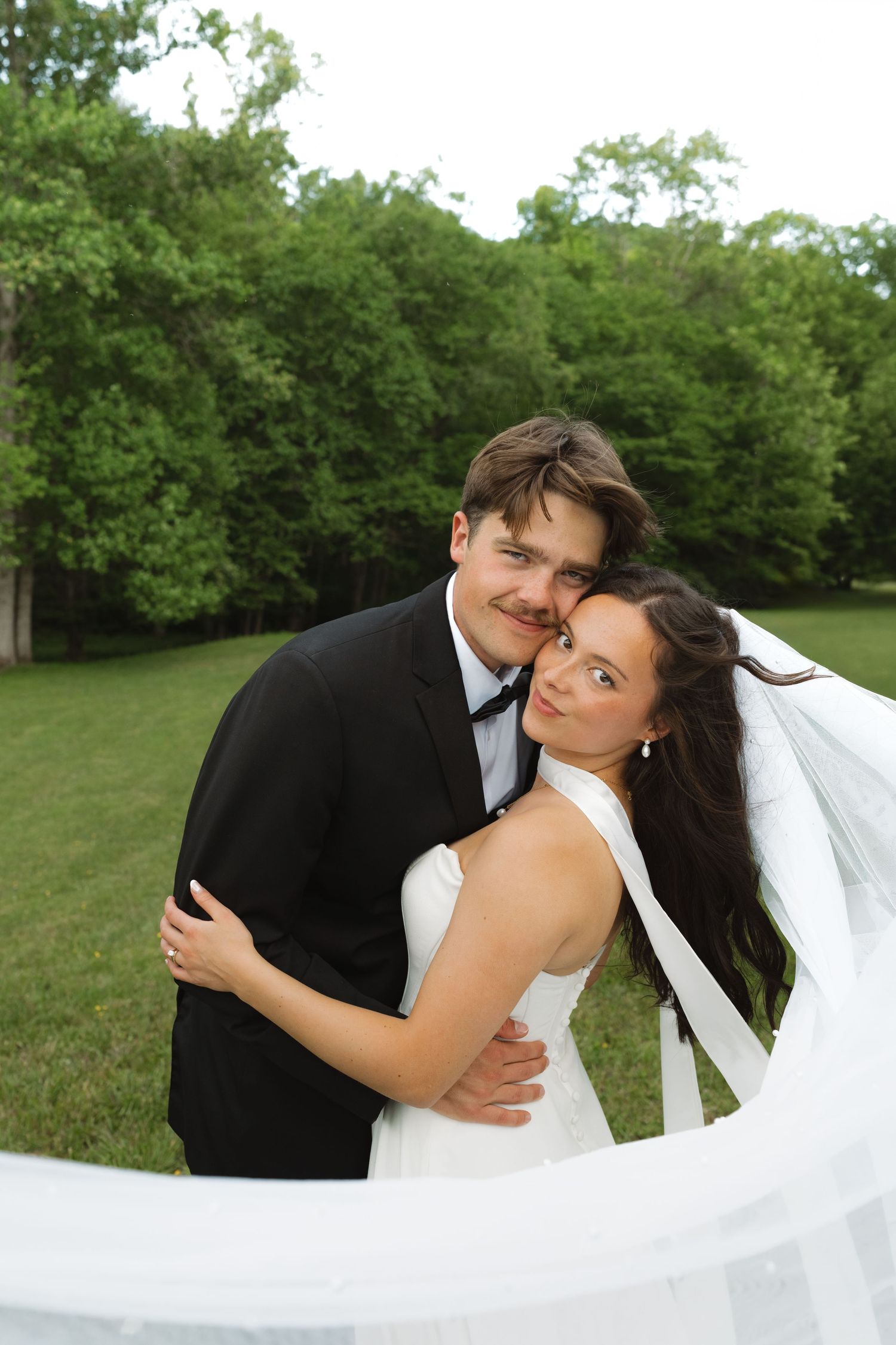 A bride and groom embrace outdoors, smiling, with green trees in the background on their wedding day.
