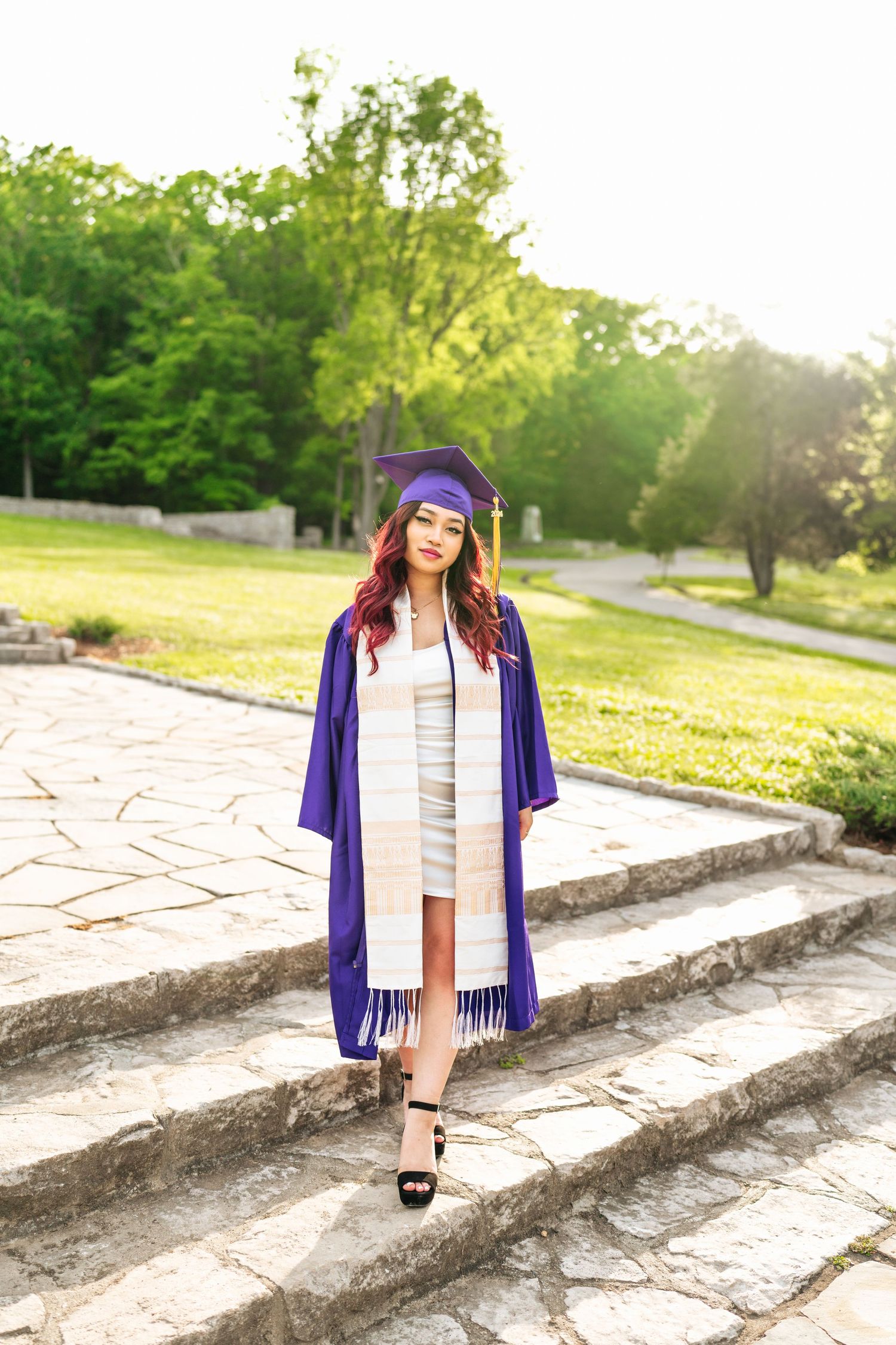 Graduate in purple cap and gown poses on stone steps during sunset photo session at Percy Warner allee.