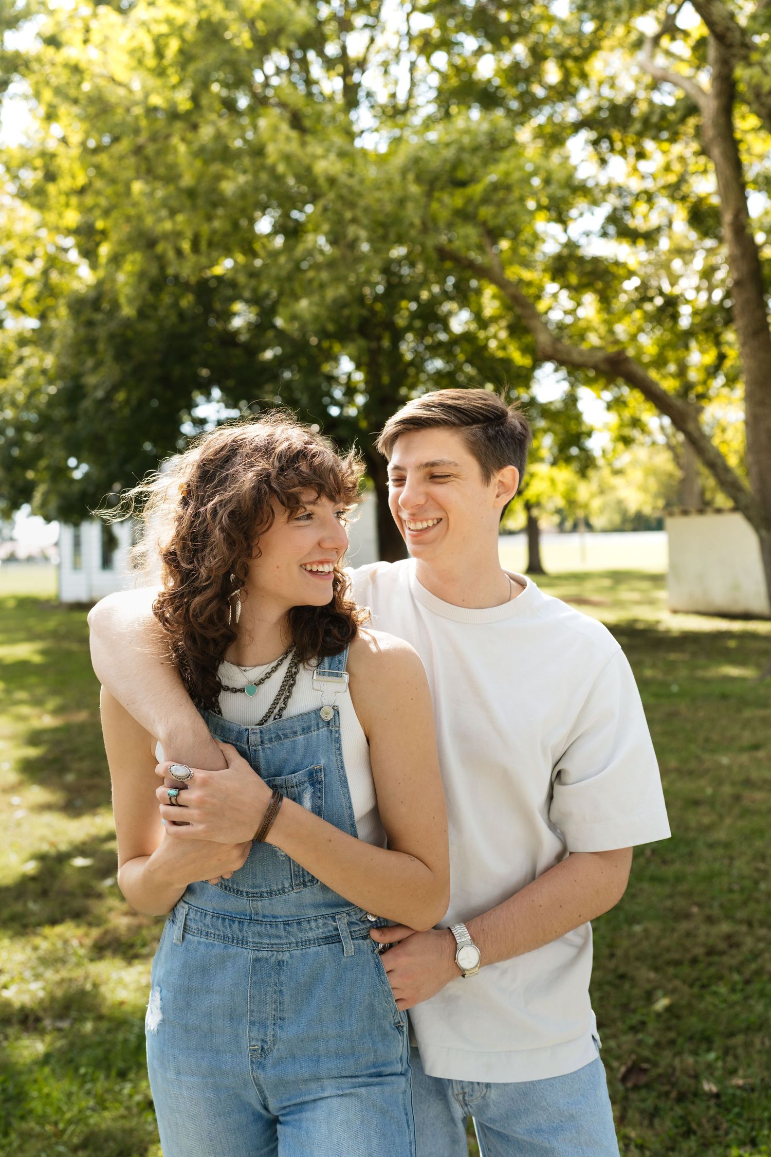 A couple shares joyful moments together in a sunlit park setting while wearing casual denim and white attire.