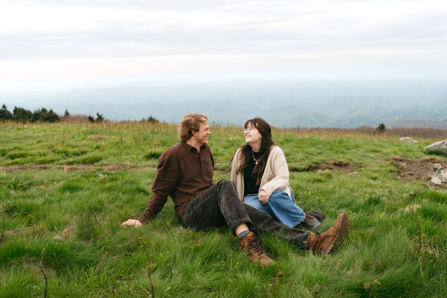 An engagement photoshoot of a couple sitting in grass on a hilltop with mountain views at Roan Mountain State Park.