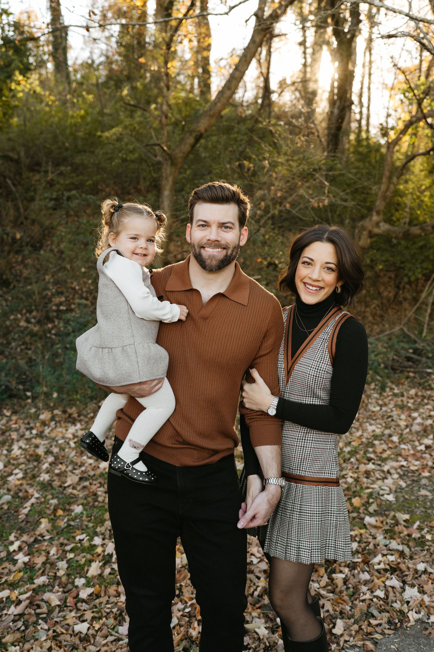 Family wearing fall sweaters and plaid outfits poses together in autumn forest setting with fallen leaves.