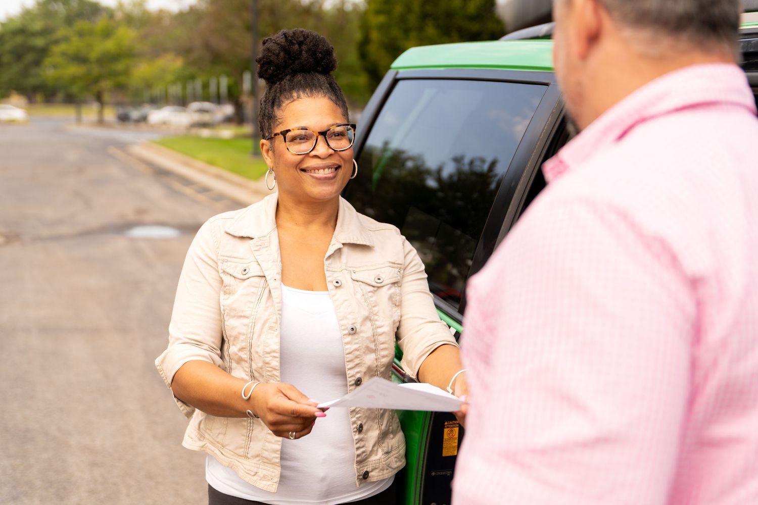 Two people exchange paperwork on a residential street next to a parked vehicle on a sunny day.