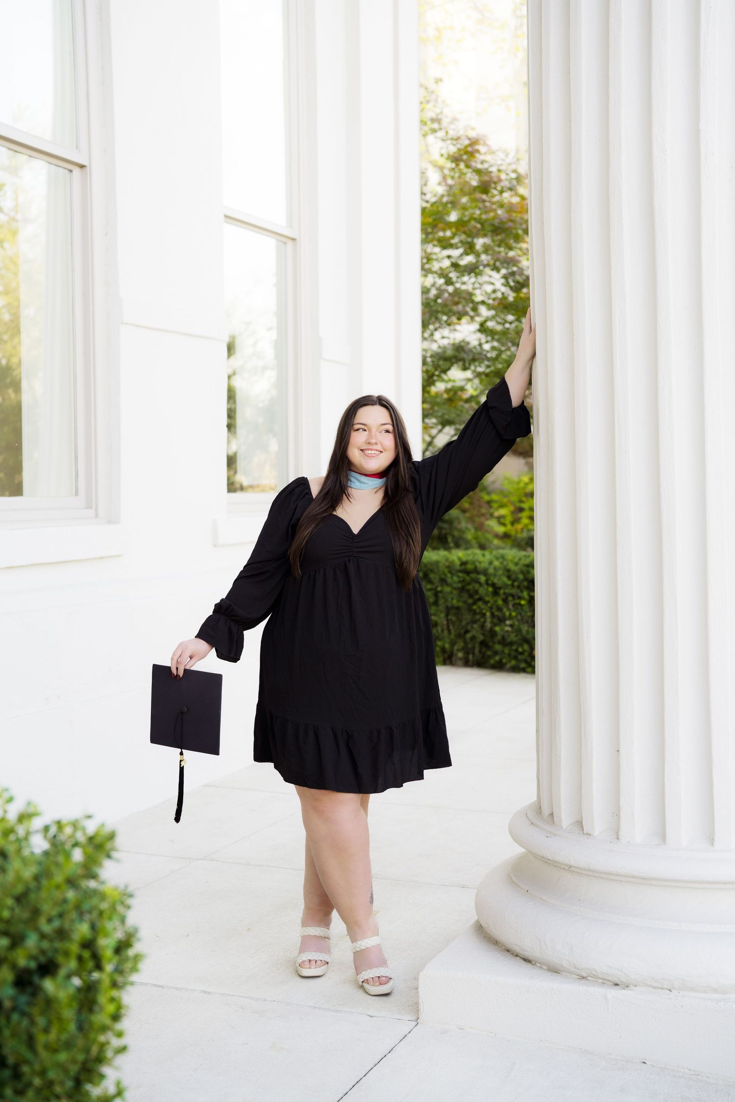 A graduate poses between white columns in a black graduation gown during a celebratory photoshoot.
