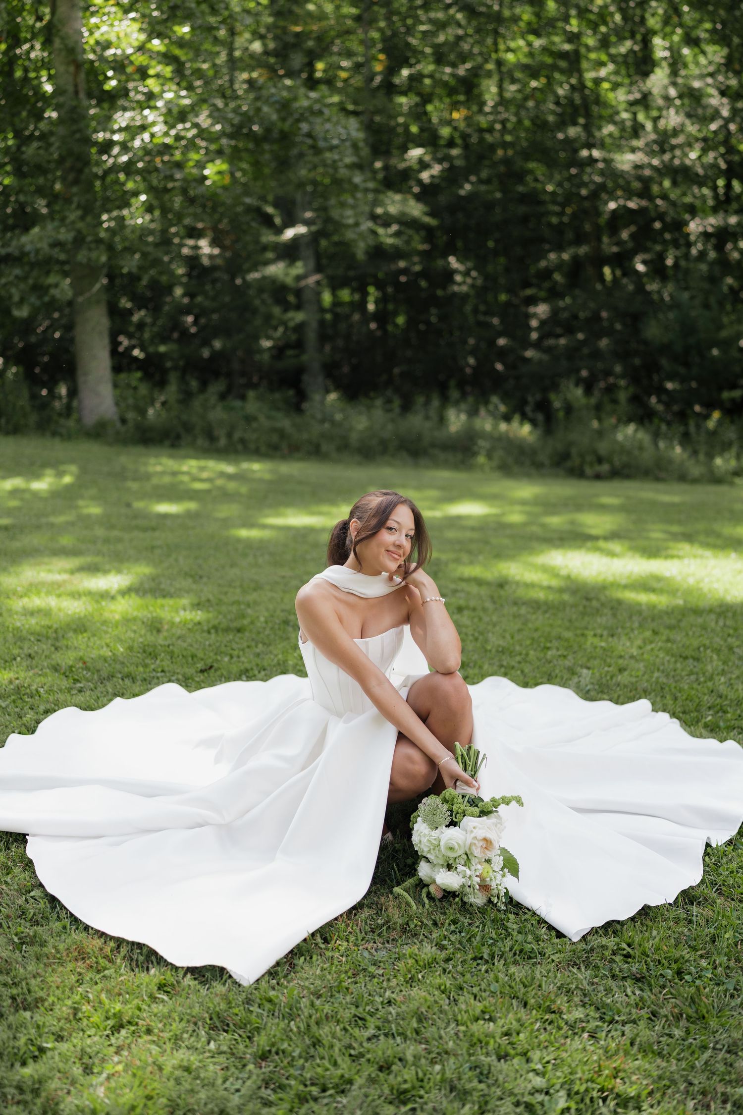 A bride in a flowing white dress lies gracefully on green grass with a bouquet during an outdoor photo session at glamping retro in erwin tn