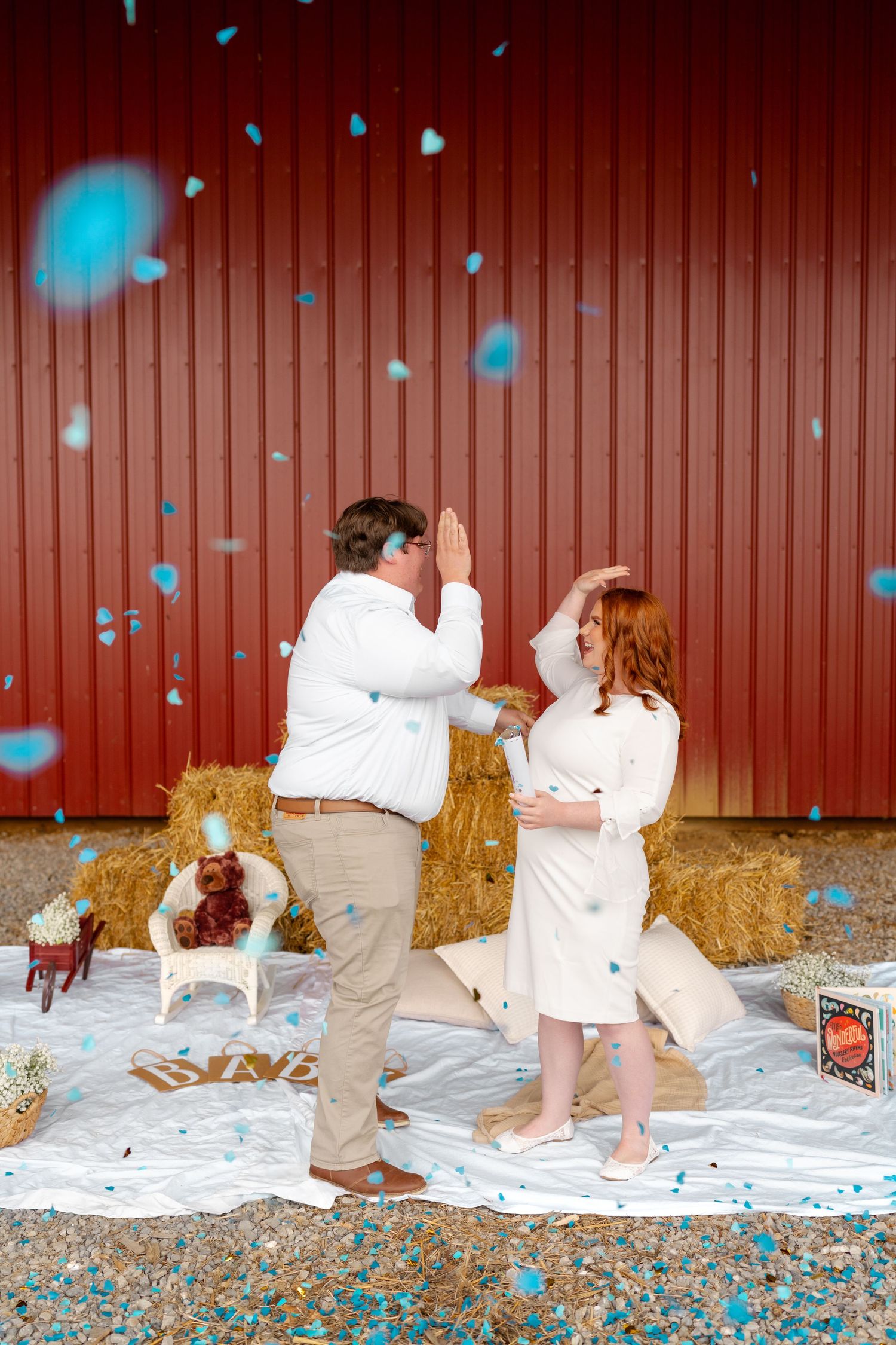 A couple in white outfits creates a playful gender reveal moment with blue confetti popping in a rustic barn setting.