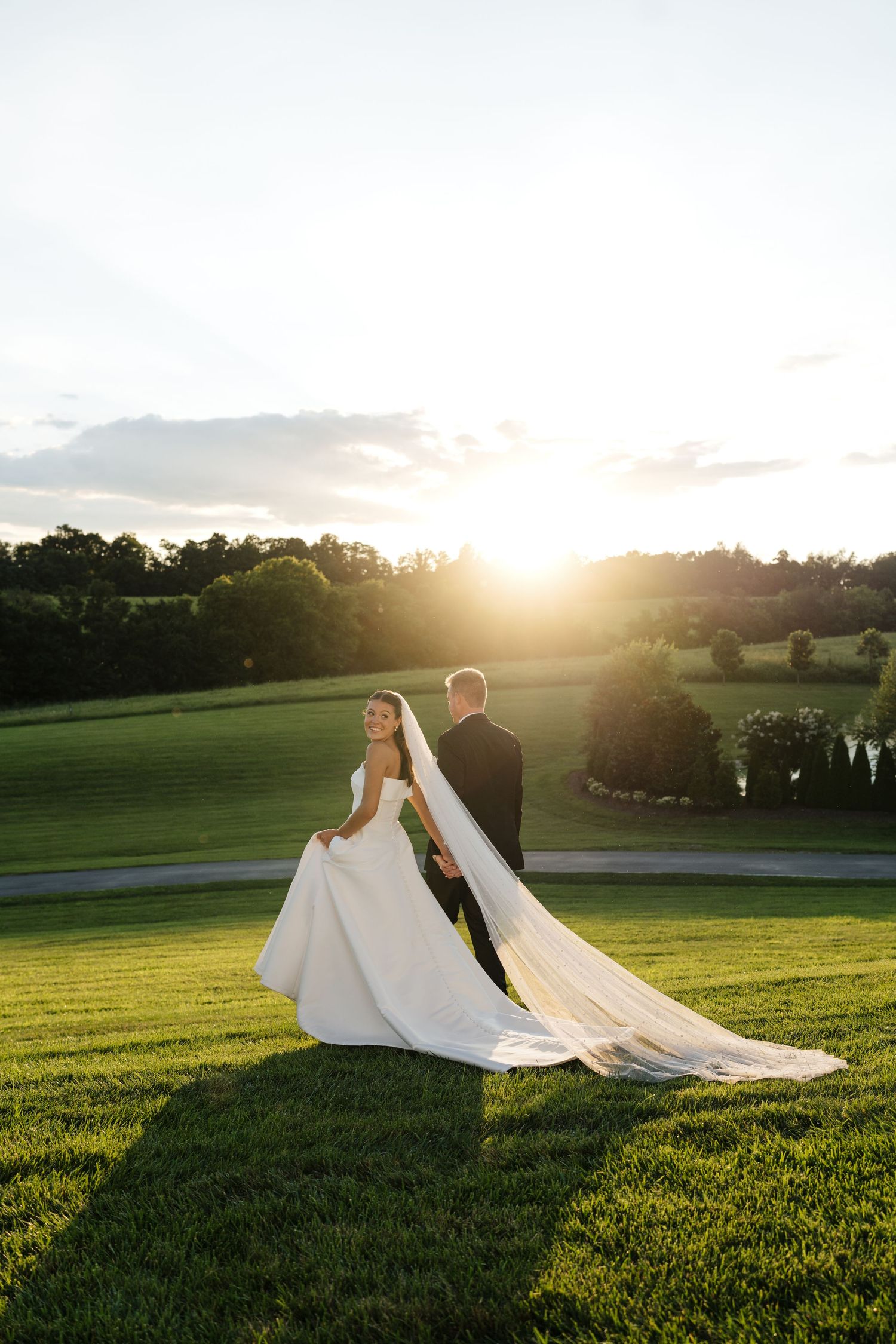 Nashville Bride and Groom walk through grassy field during golden hour with flowing white dress train at White Dove Barn in Beechgrove, TN.