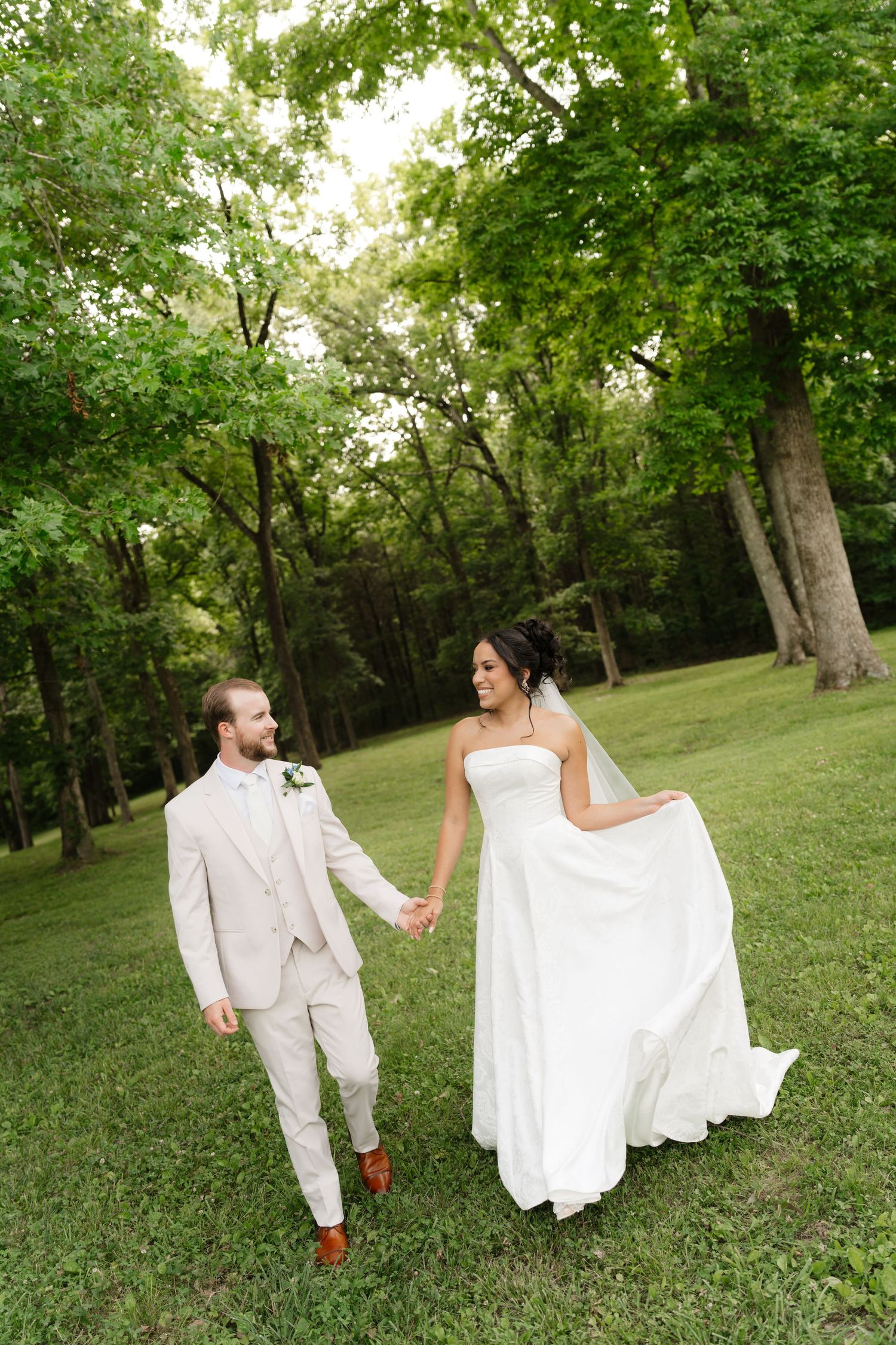 A bride in a white gown and groom in a tan suit hold hands while walking through a lush green outdoor setting.