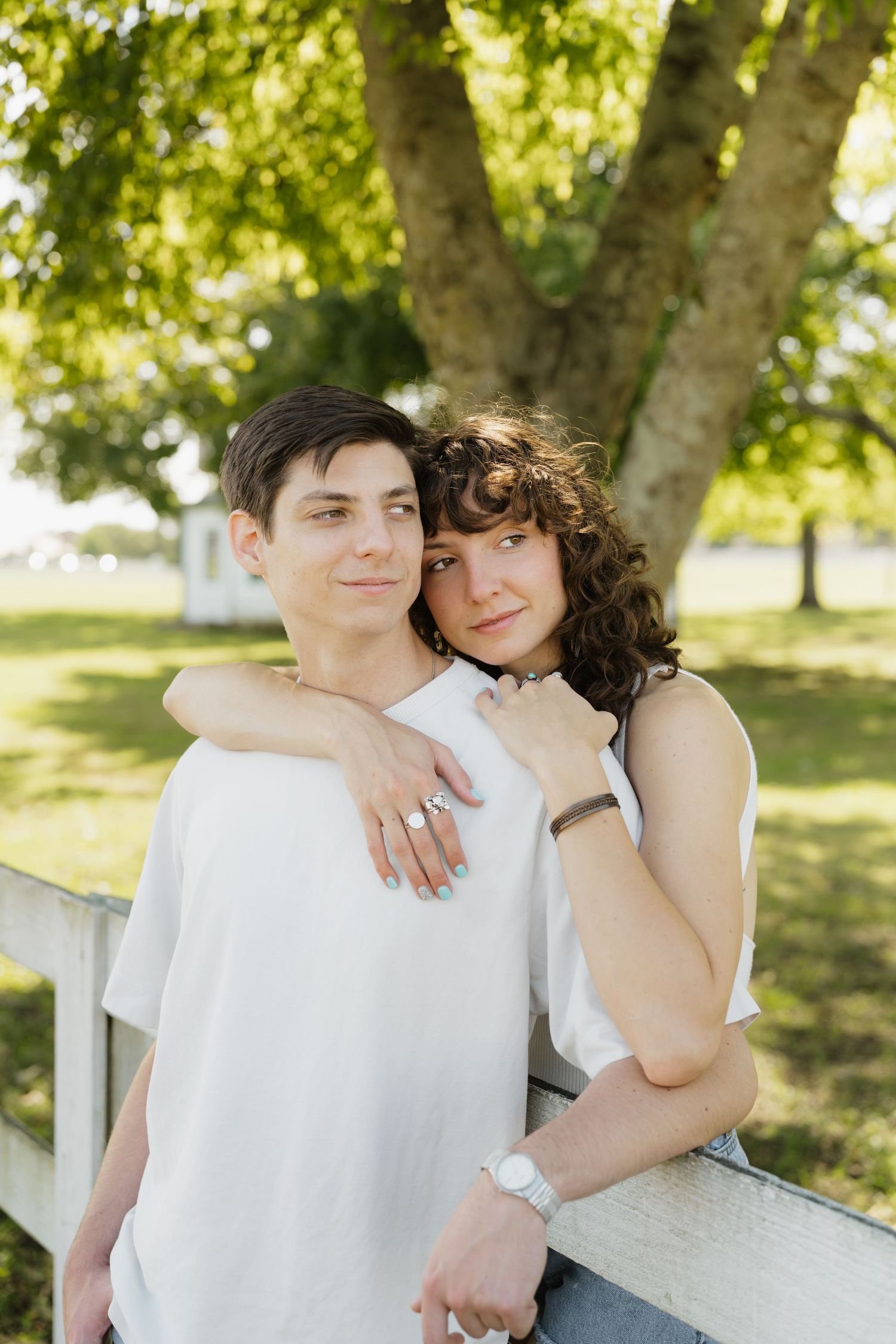 A couple in white outfits embraces lovingly on a sunny day in a park with green trees in the background.