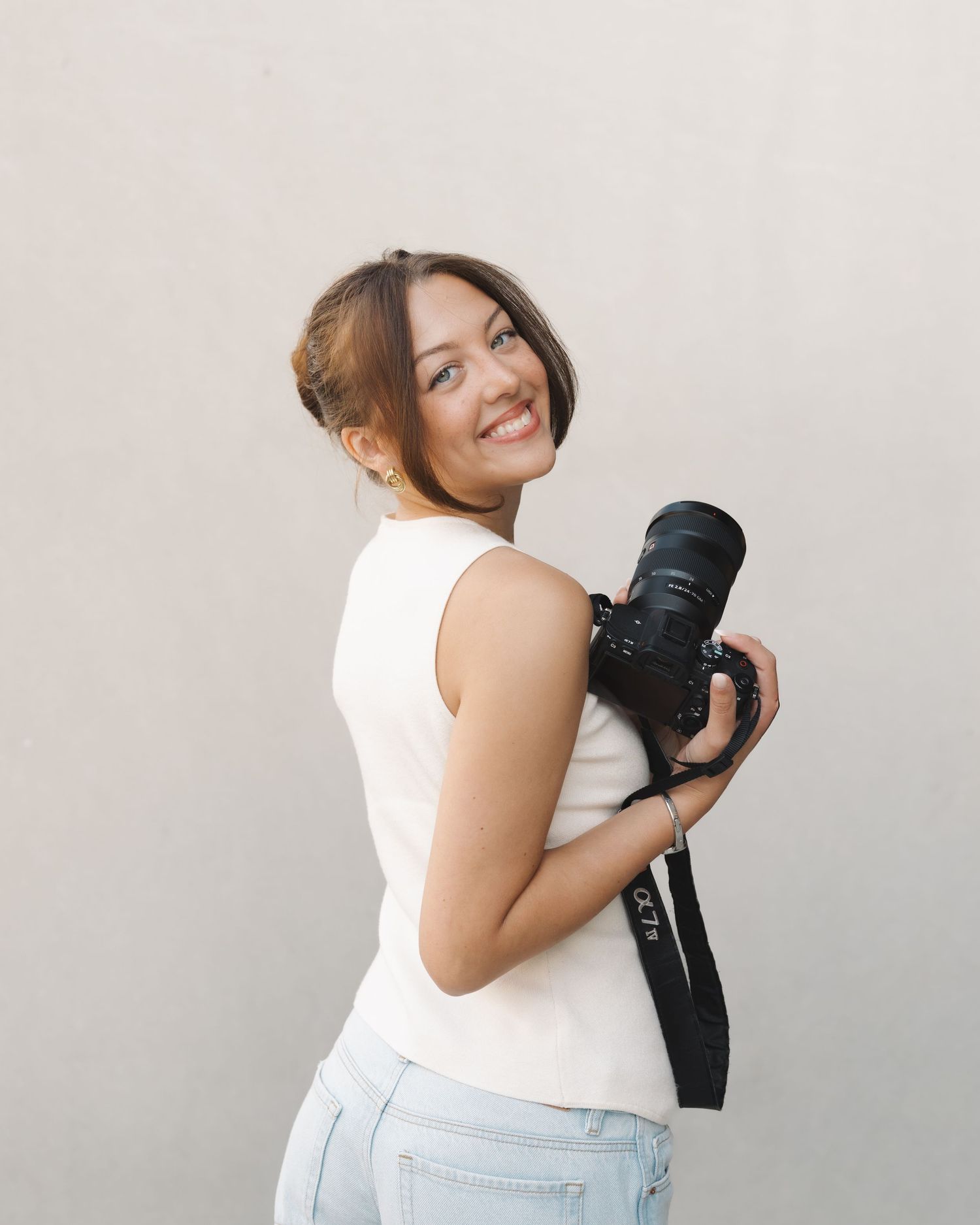 Photographer, Chloe Hughes in white top and jeans holds professional camera against neutral background.