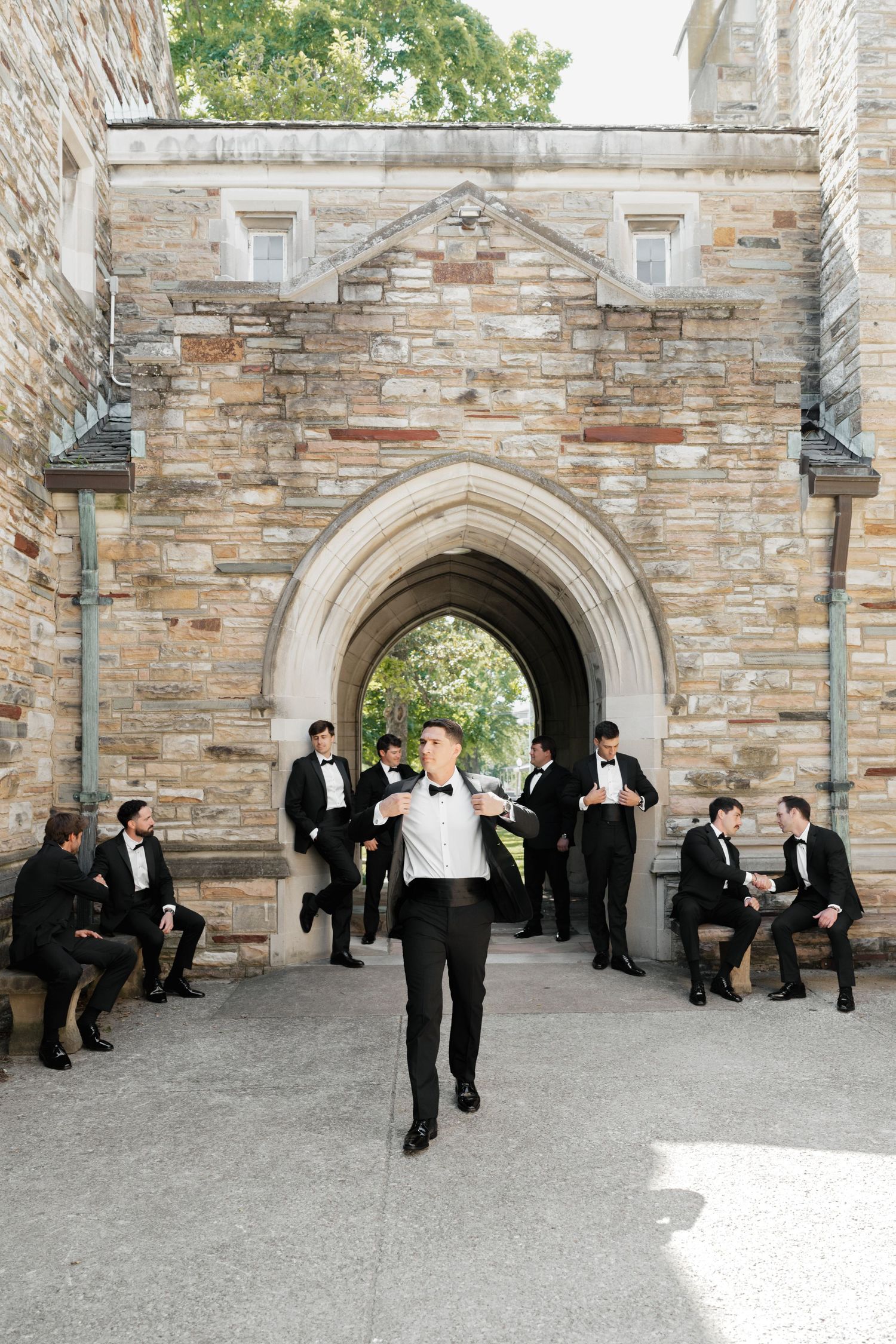 Groom adjusting bow tie walks toward camera with groomsmen in black tuxedos relaxing near stone archway.
