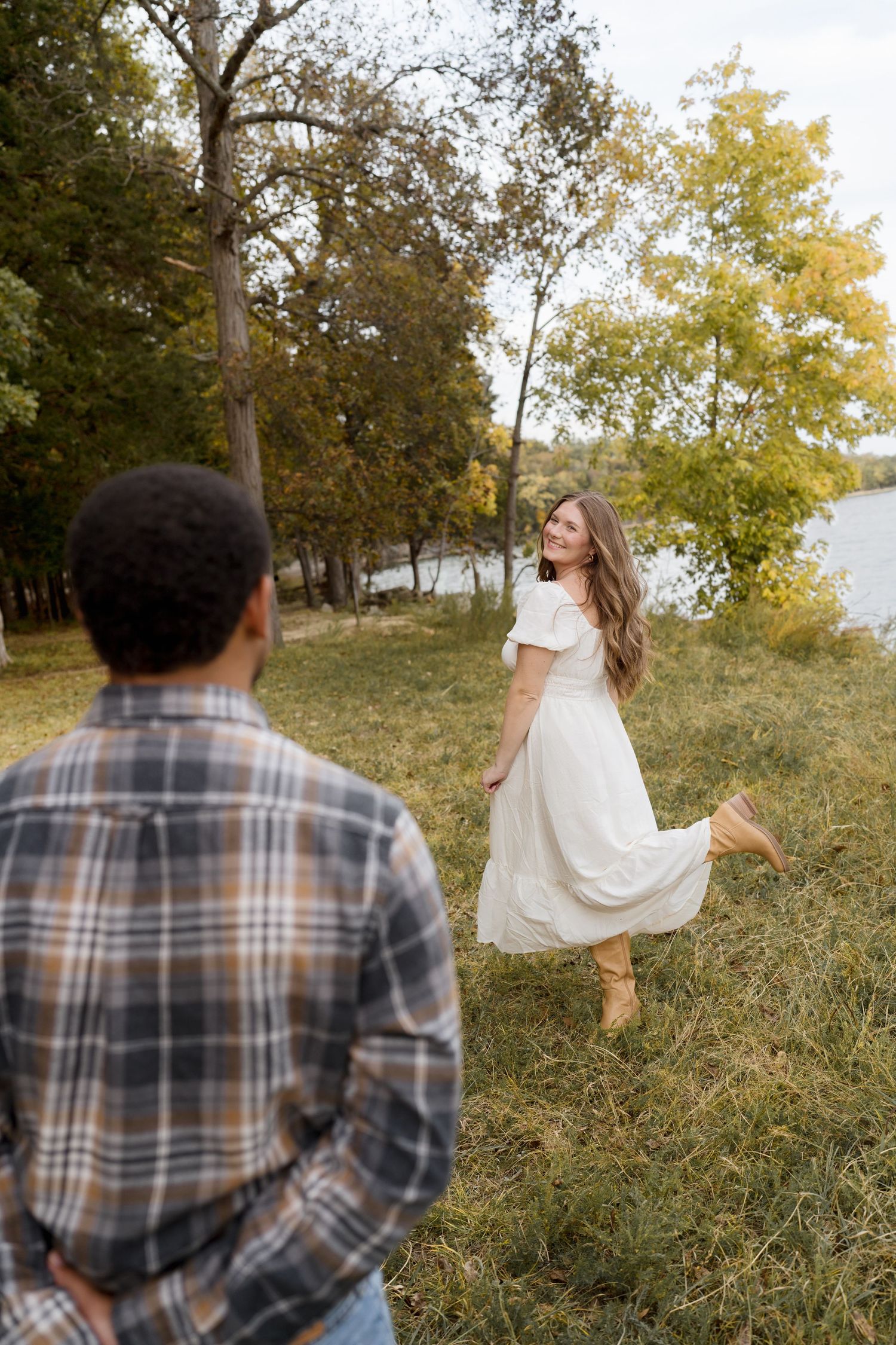 A person in a white dress twirls playfully in a grassy field during golden hour while someone in plaid watches.