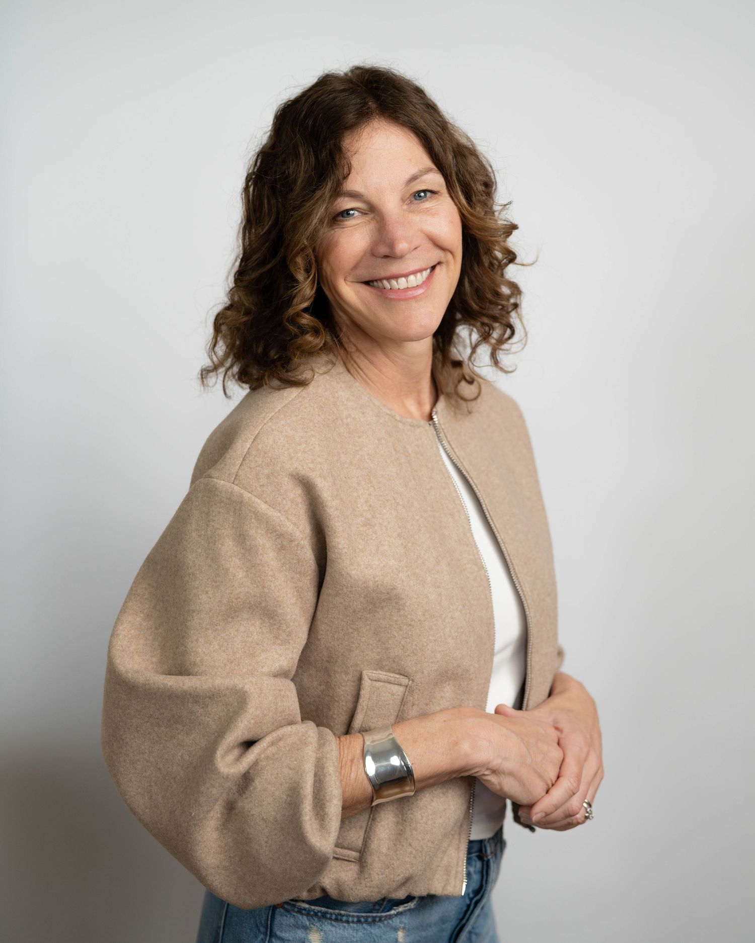 A person wearing a beige sweater and jeans poses joyfully against a white backdrop in a studio setting.