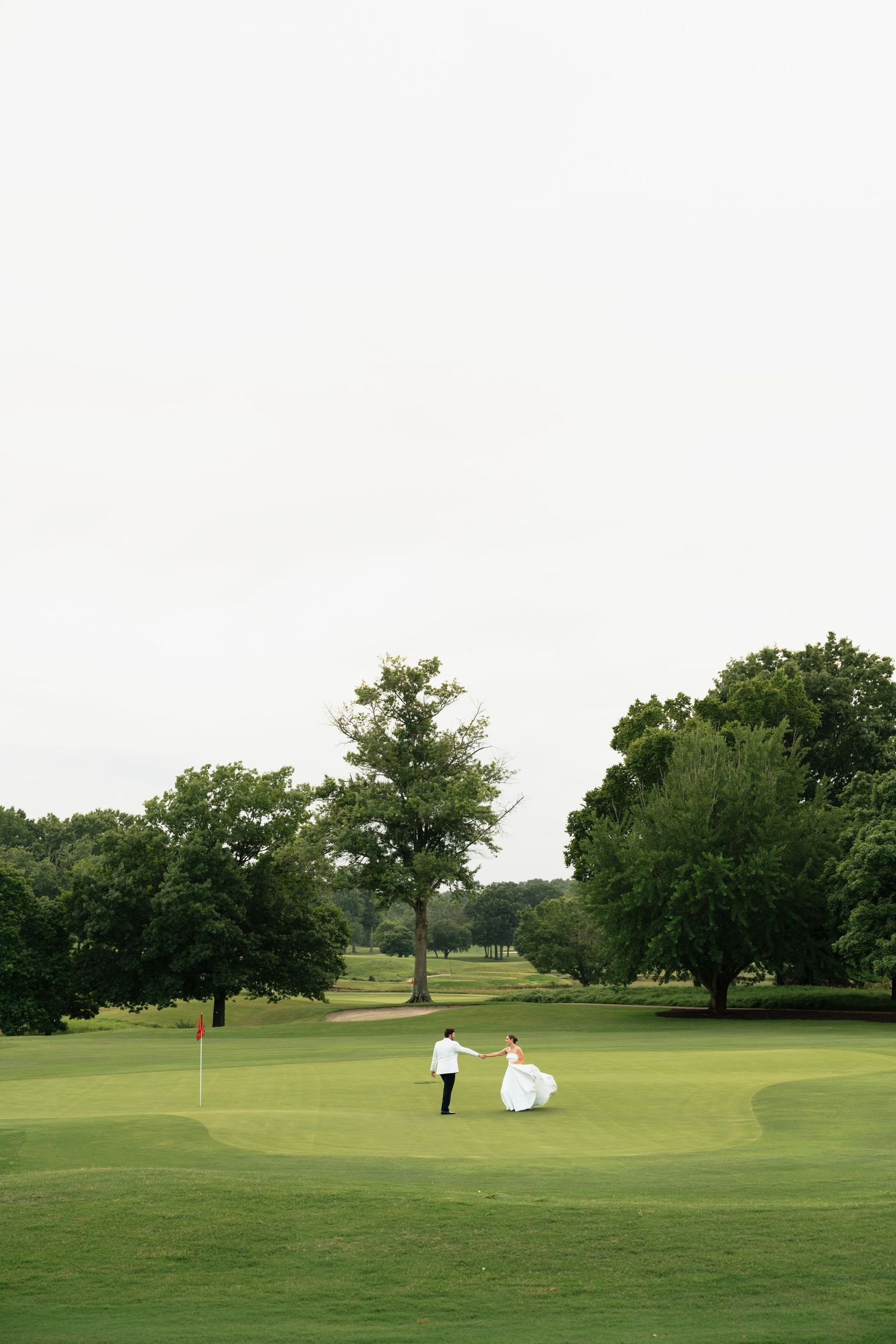 Bride and groom holding hands on a lush green golf course surrounded by tall trees on their wedding day.