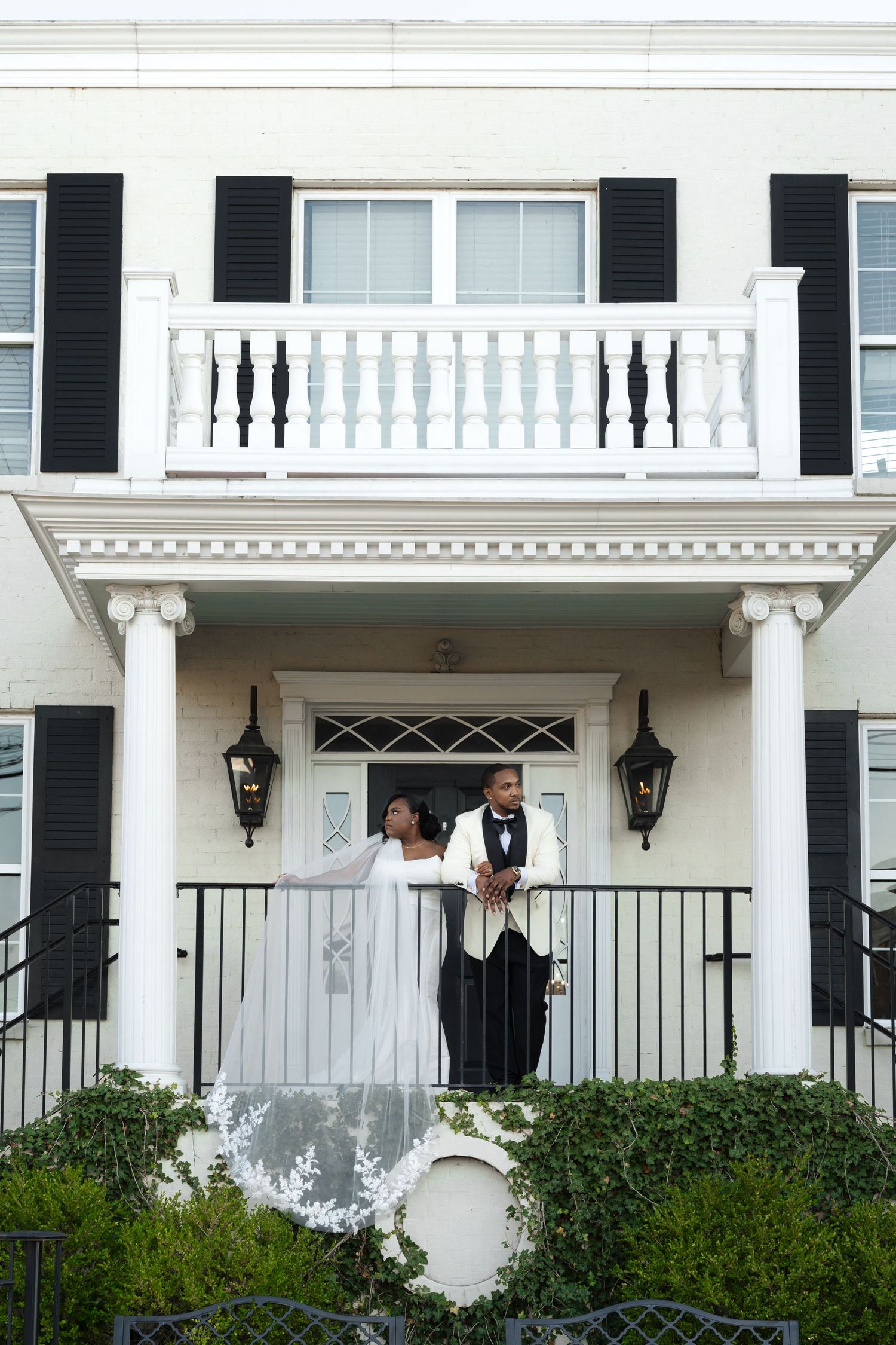 Bride and groom in formal wedding attire pose on the porch of an elegant white colonial mansion.
