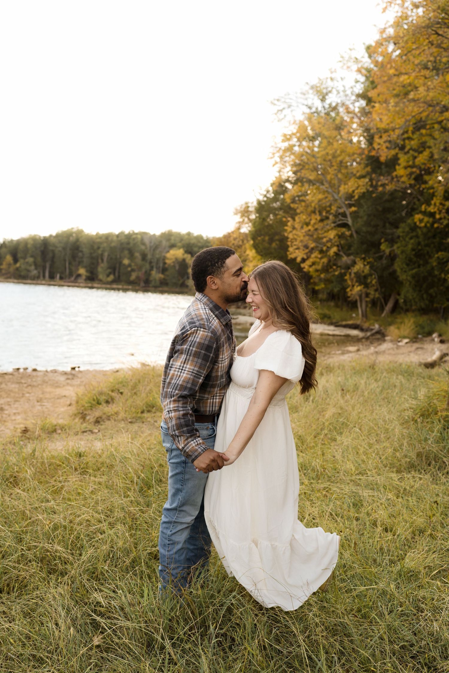 A couple shares a romantic moment in a scenic fall setting by a lake with golden autumn foliage in the background.