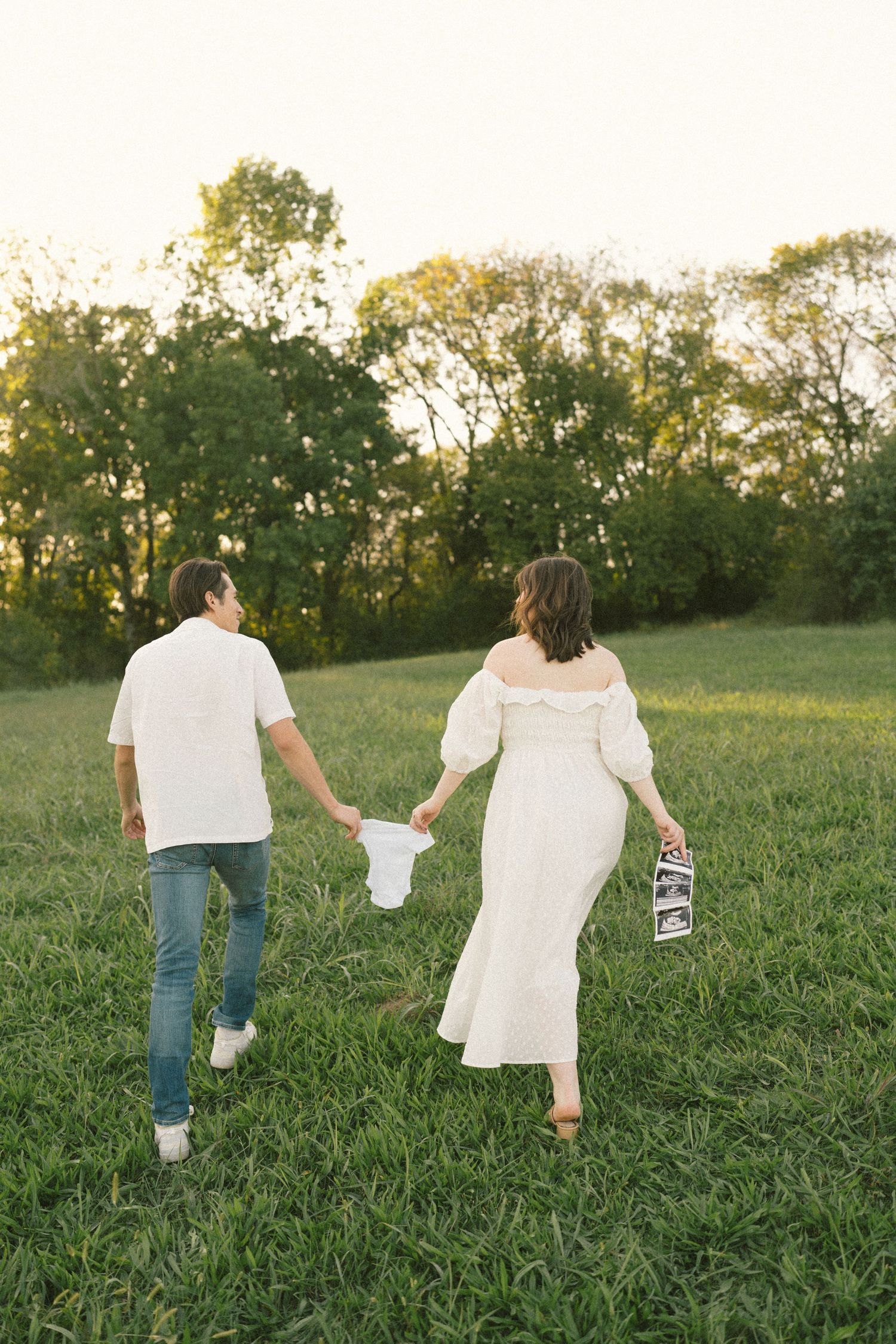 Mom and Dad walking through grassy field at sunset holding a onesie to announce pregnancy.