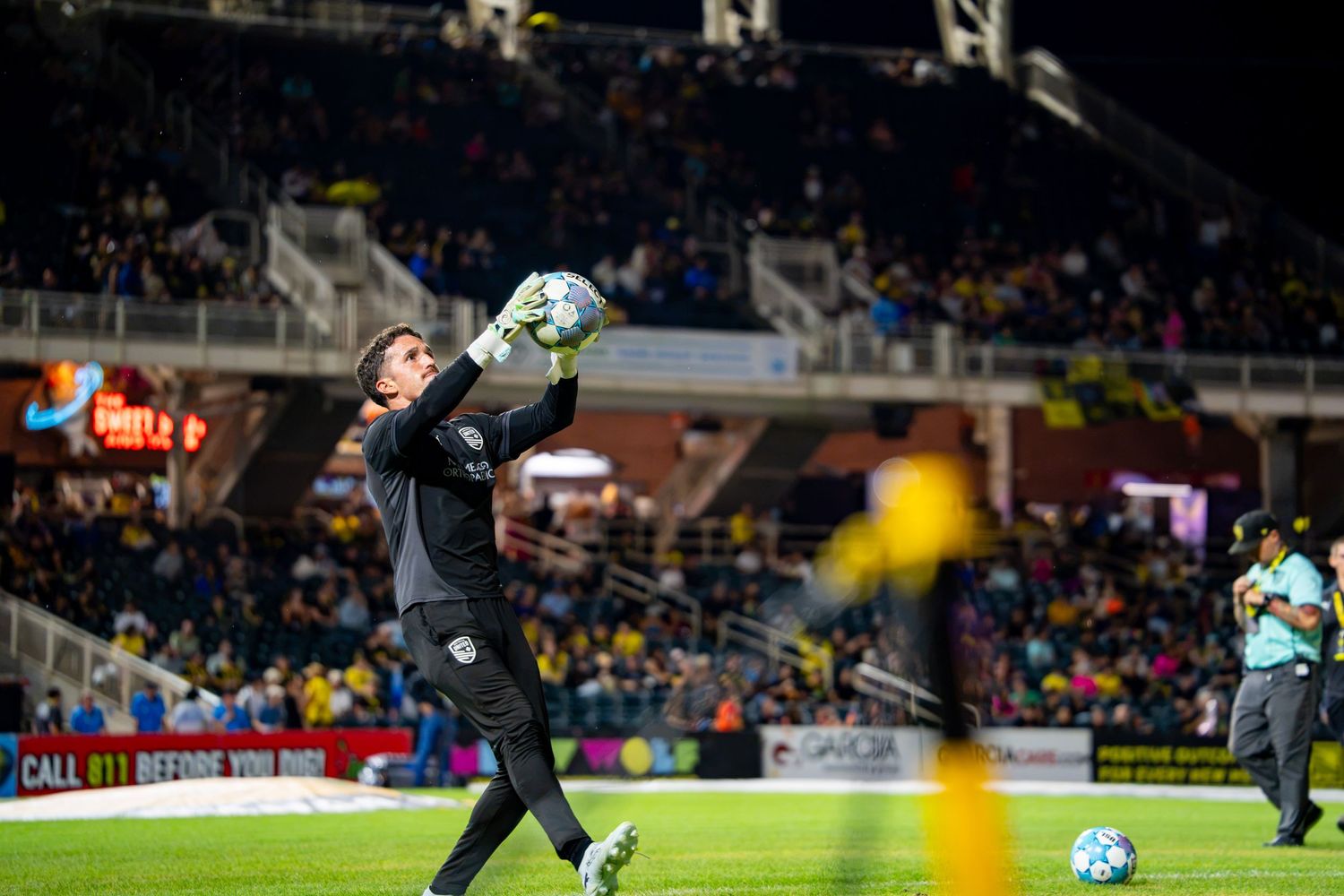 Soccer goalkeeper catches ball during night match at stadium with yellow and black team colors in background.