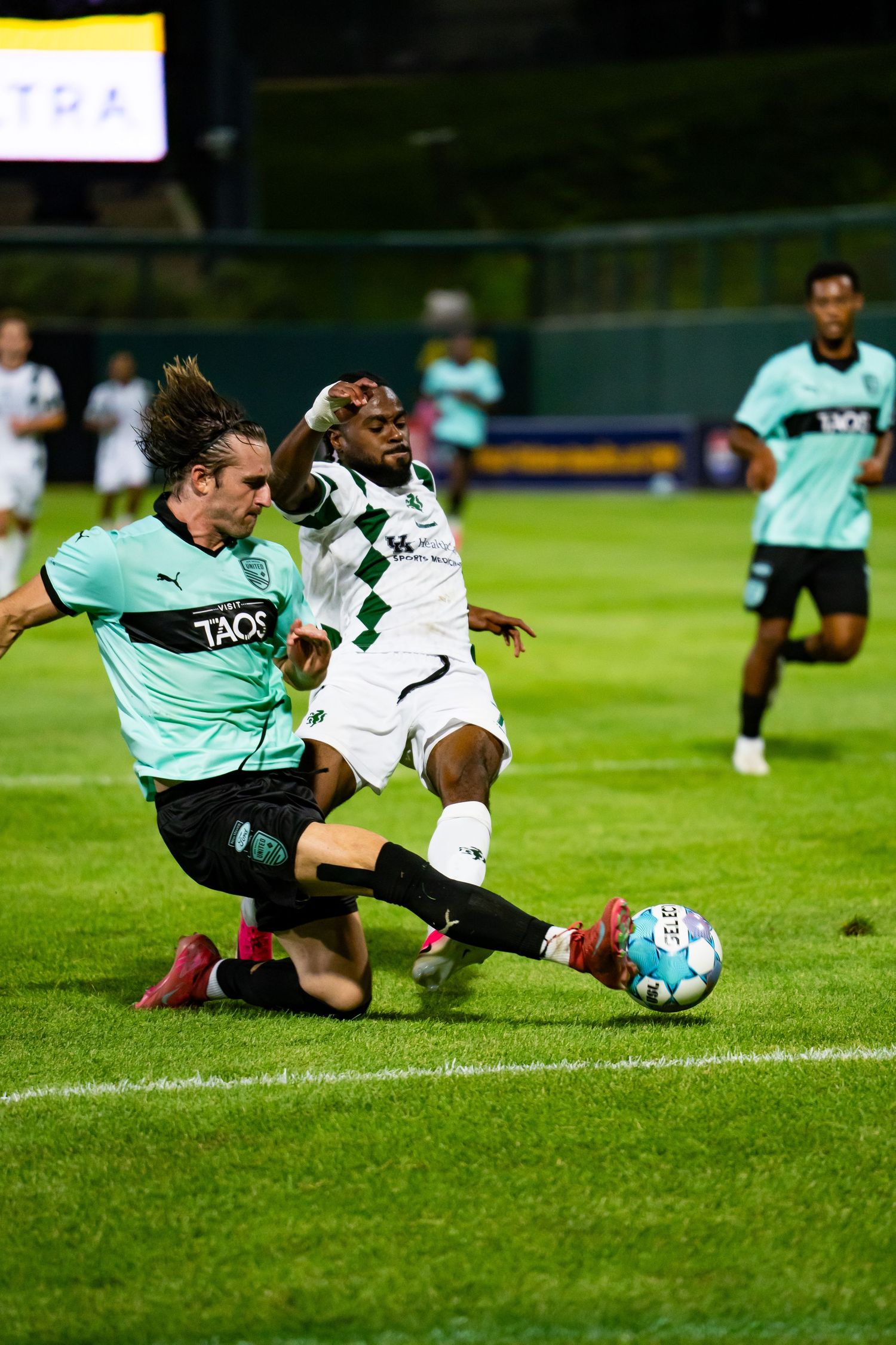Two soccer players compete for the ball during a night match, one sliding in a tackle on a green field.