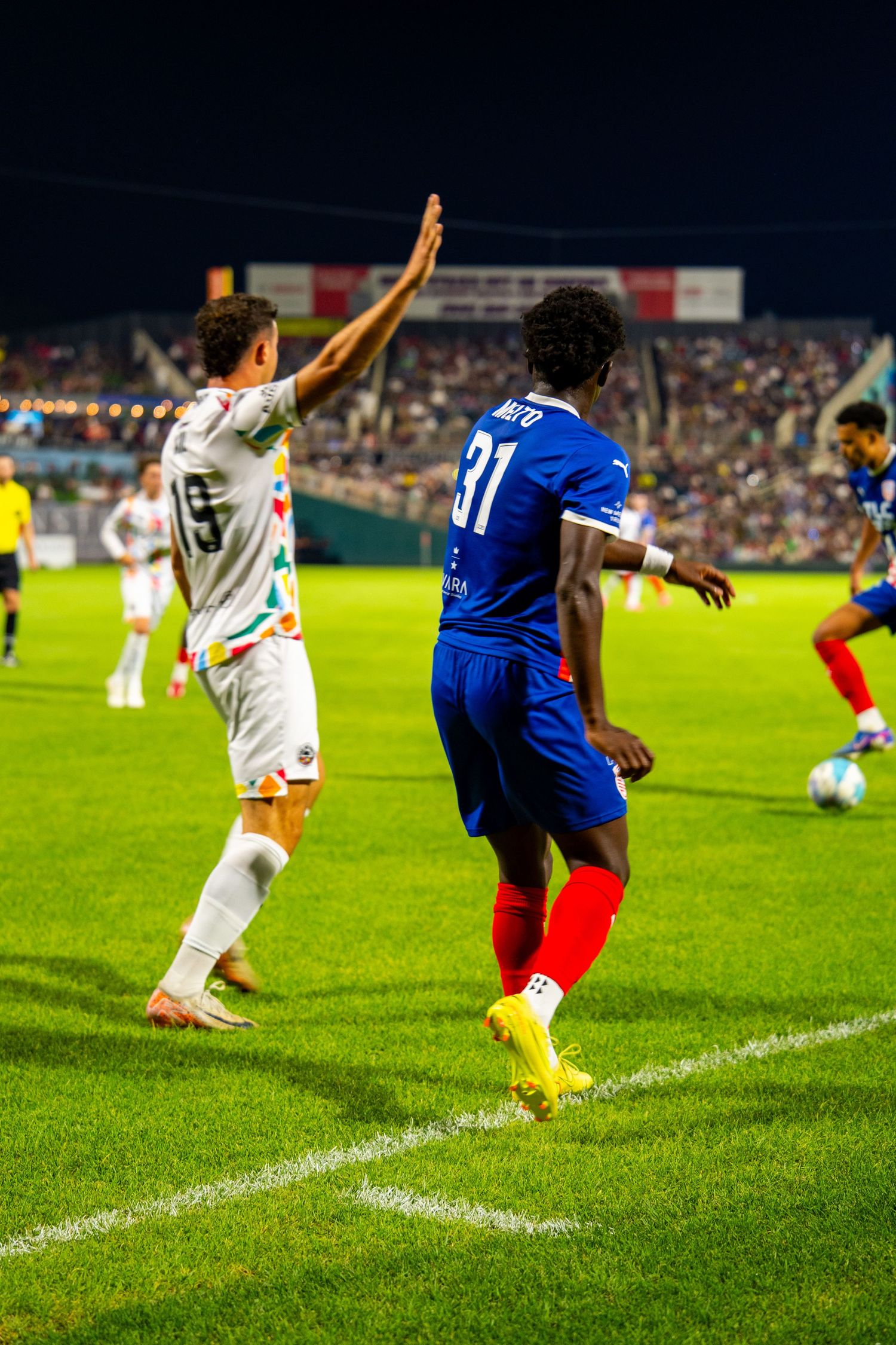Soccer players in action at night game, player #31 in blue jersey faces opponent in white uniform on green field.