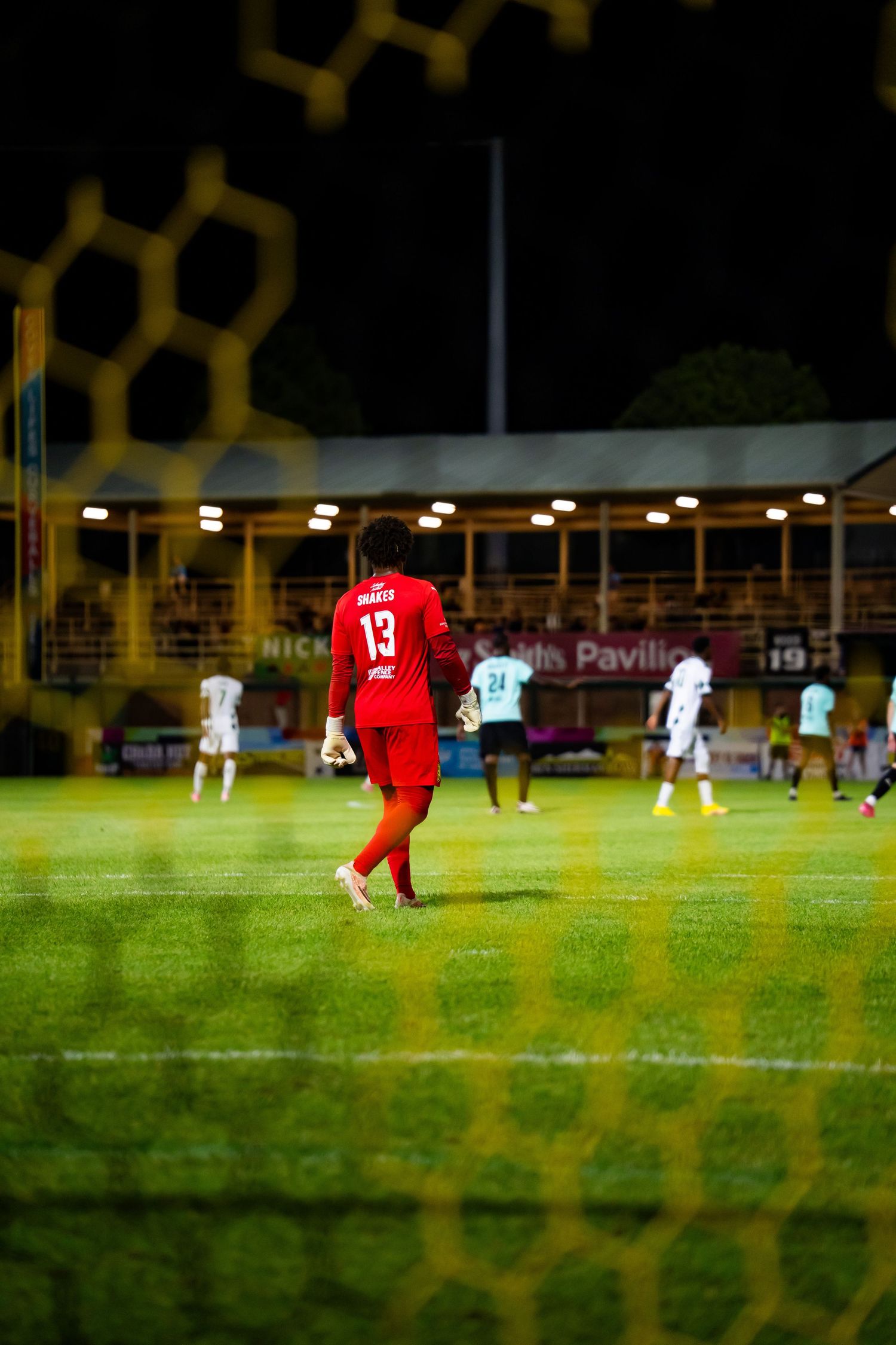 Soccer player wearing red jersey number 13 walks on illuminated green field during night match viewed through goal net.