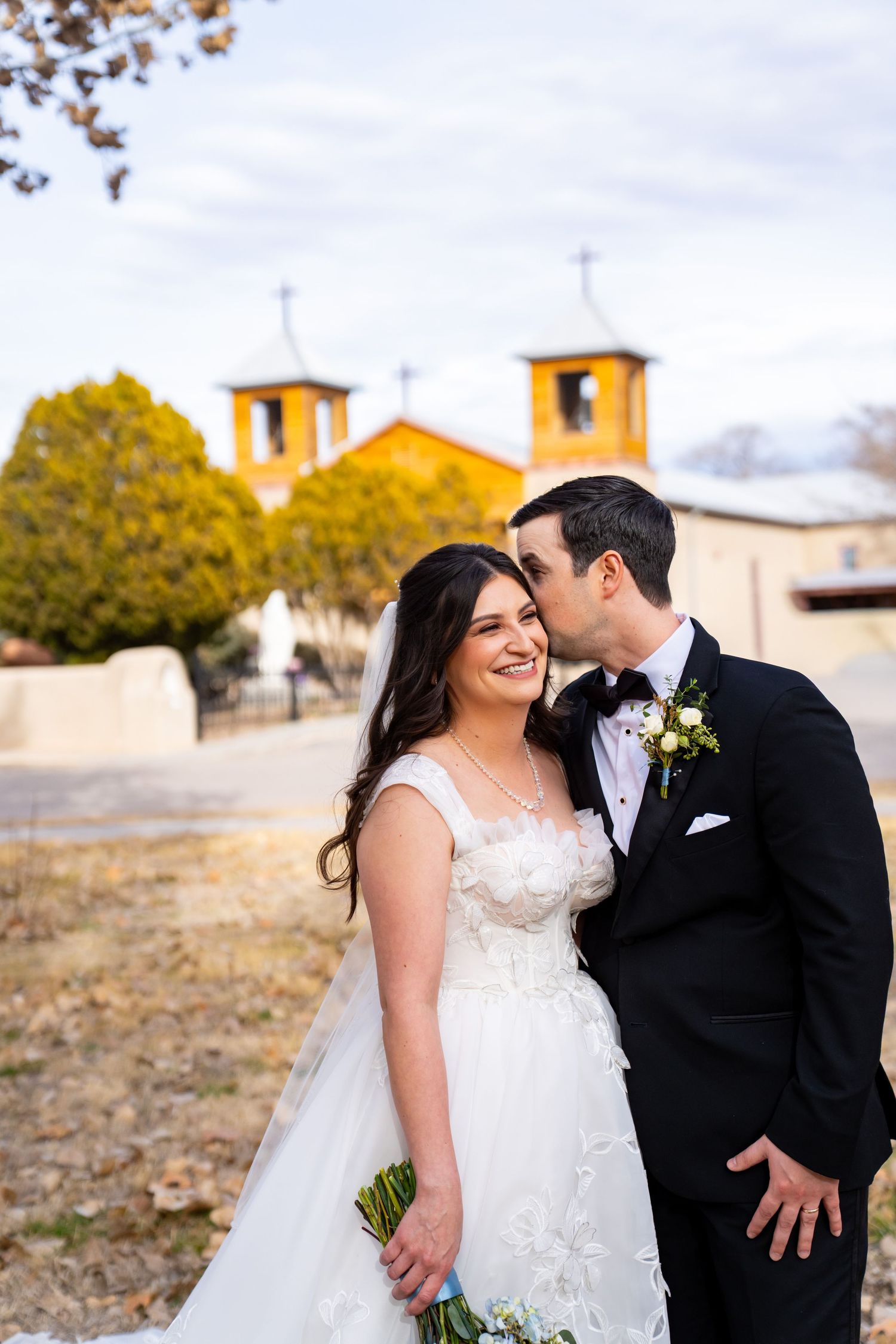Bride and groom share a romantic kiss outside a historic adobe church with yellow bell towers on their wedding day.