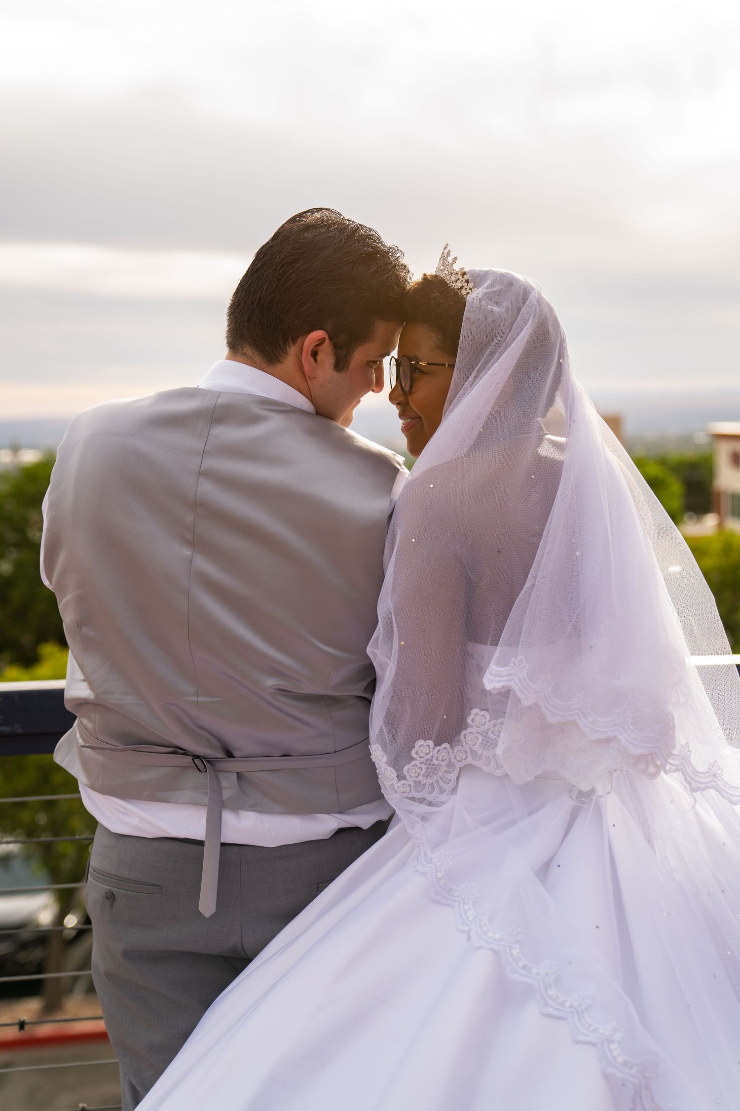 A romantic wedding couple embraces on a balcony during sunset, with the bride wearing a flowing white gown and veil.