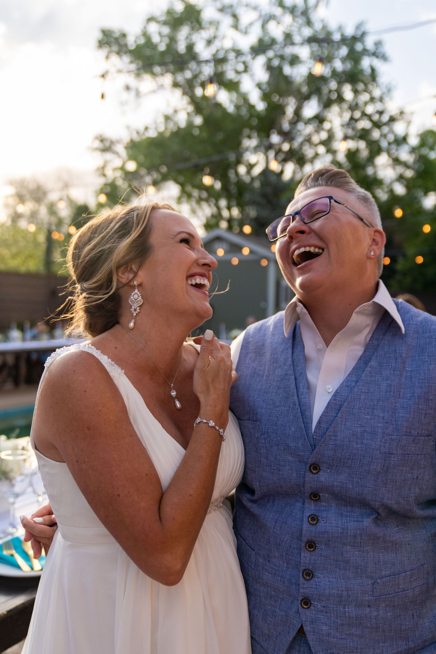 A joyful couple shares a candid laughing moment at an outdoor wedding reception with string lights in the background.