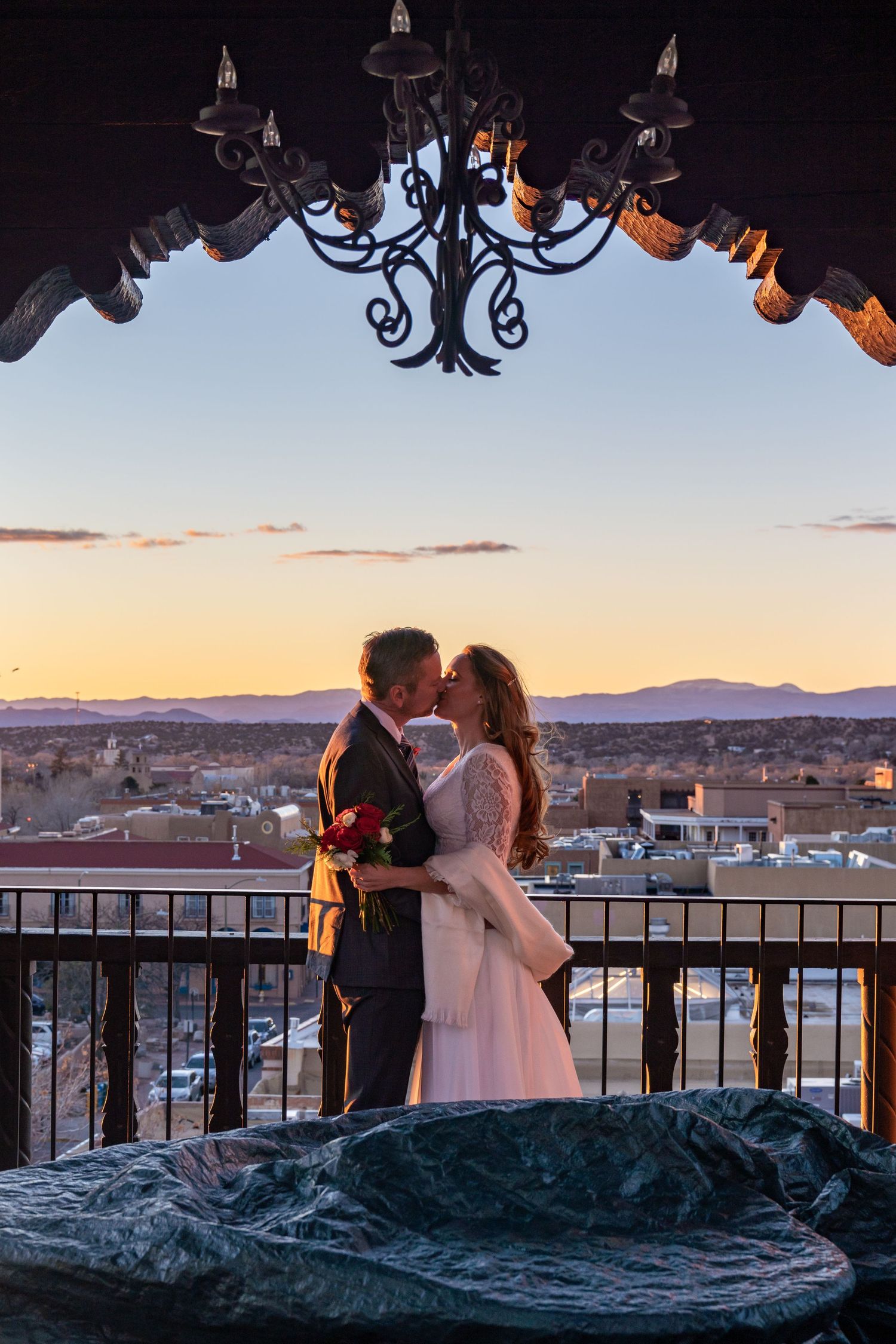 A romantic sunset wedding moment on a balcony with an ornate chandelier and cityscape view in the background.