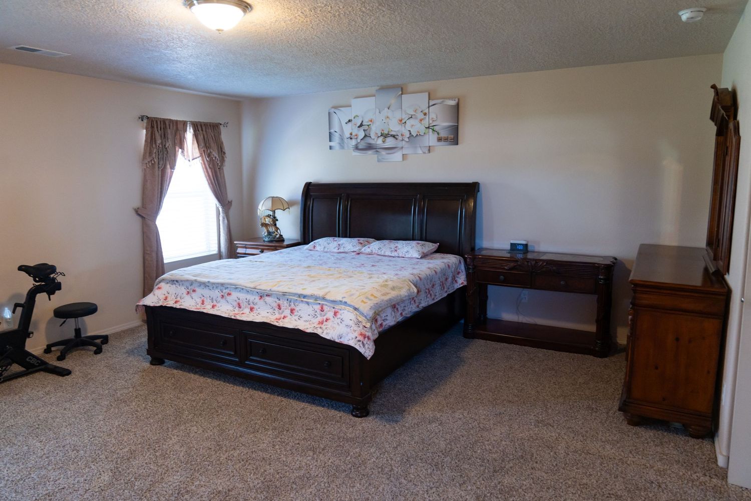A spacious bedroom with dark wood furniture, beige carpet, and floral bedding features an exercise bike in the corner.