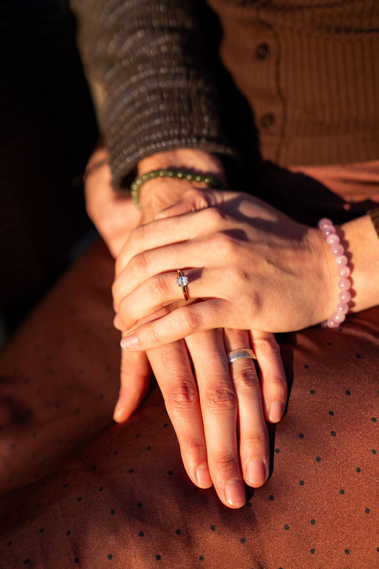 Two hands resting together showing an engagement ring and wedding band, with beaded bracelets, in warm sunlight.