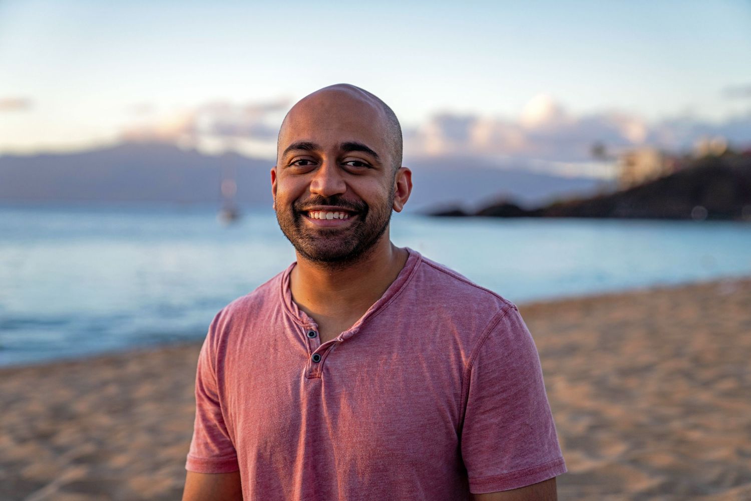 Portrait of person smiling warmly on beach at sunset wearing pink shirt.