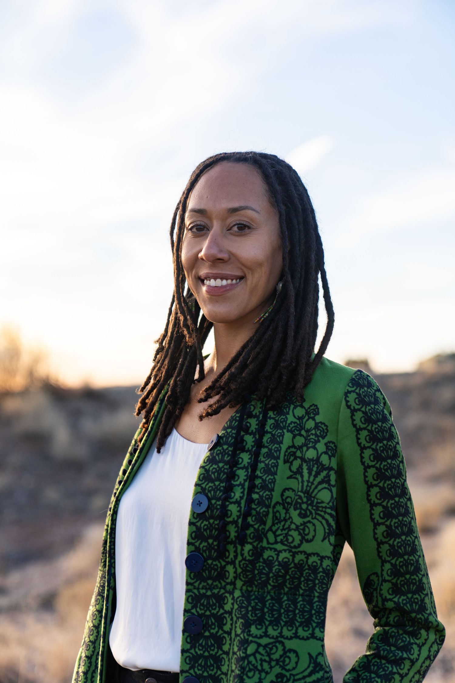 Person in ornate green jacket smiling against desert landscape background.