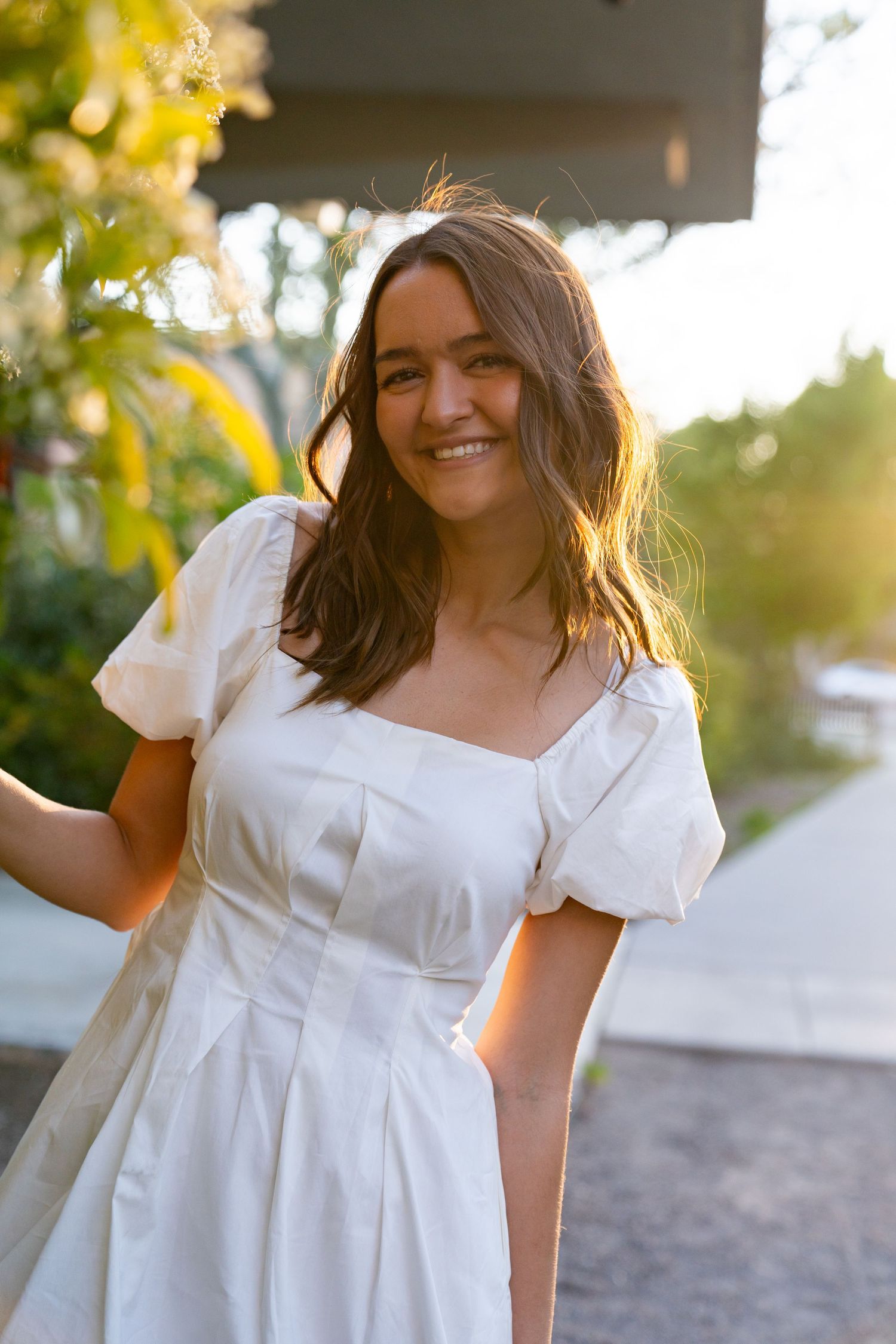 Woman in white summer dress poses outdoors during golden hour sunset.