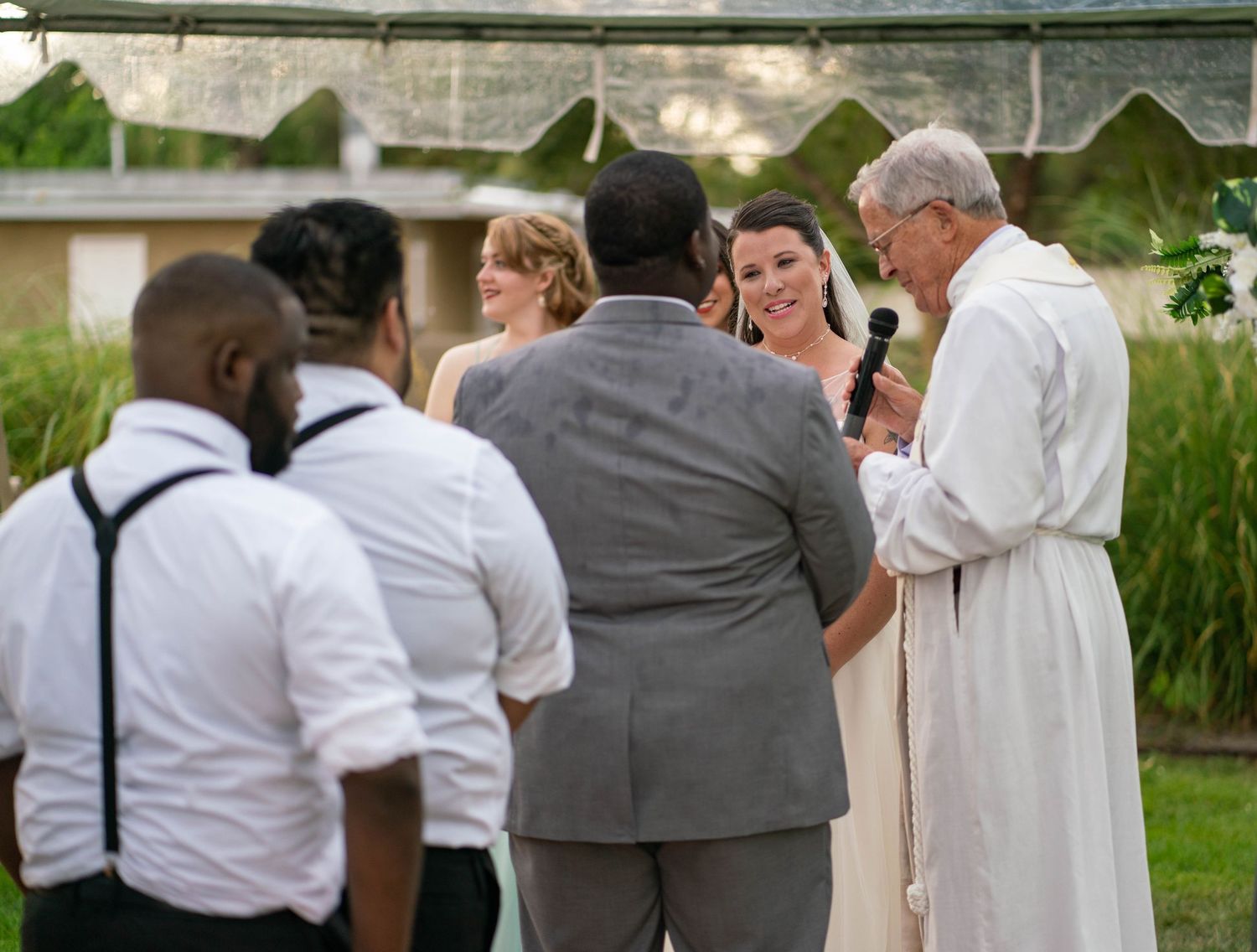 Wedding party in formal attire gathers outdoors in a garden setting with white and gray color scheme.
