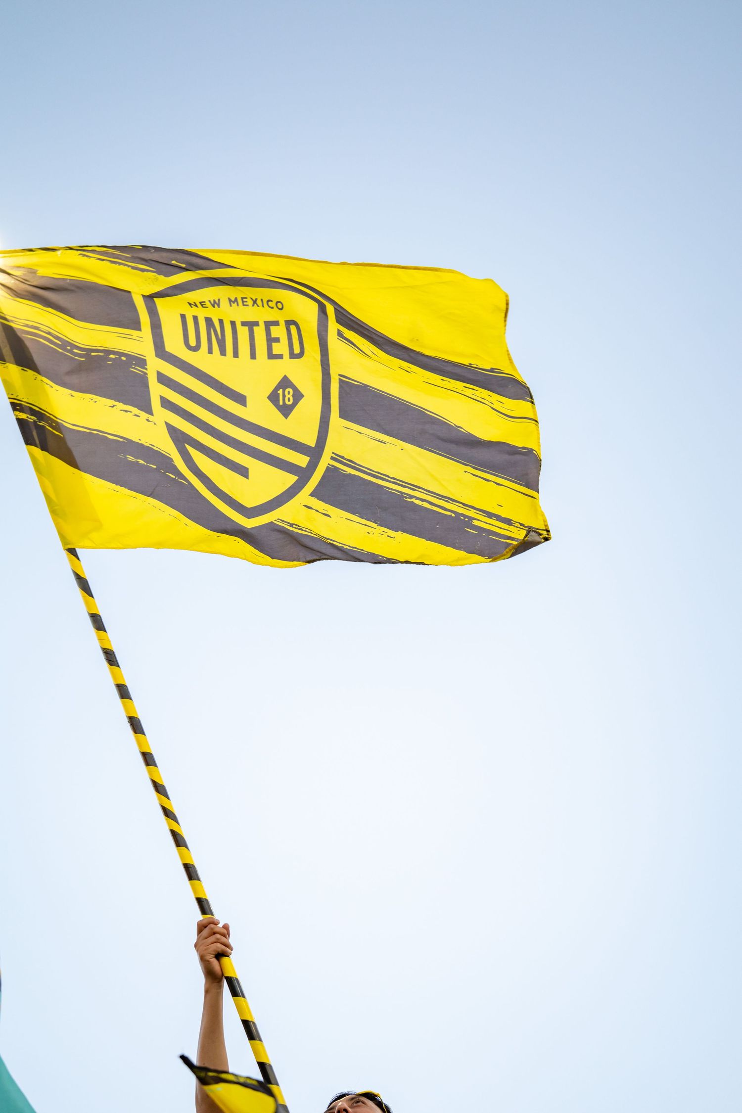 A New Mexico United soccer fan waves a yellow and gray team flag against a clear blue sky.