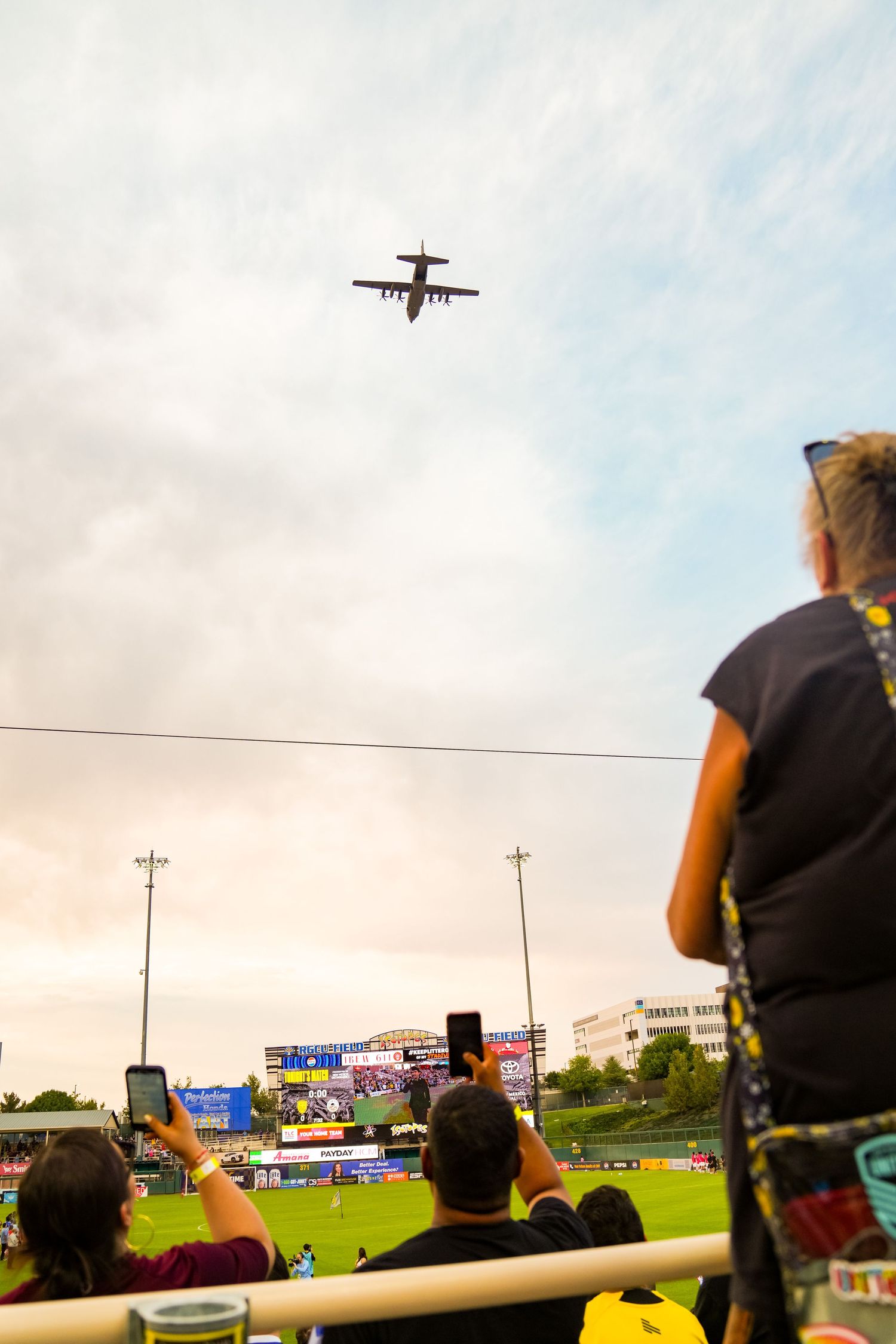 Crowd at baseball stadium watches military cargo plane fly overhead, fans photographing the flyover against cloudy sky.