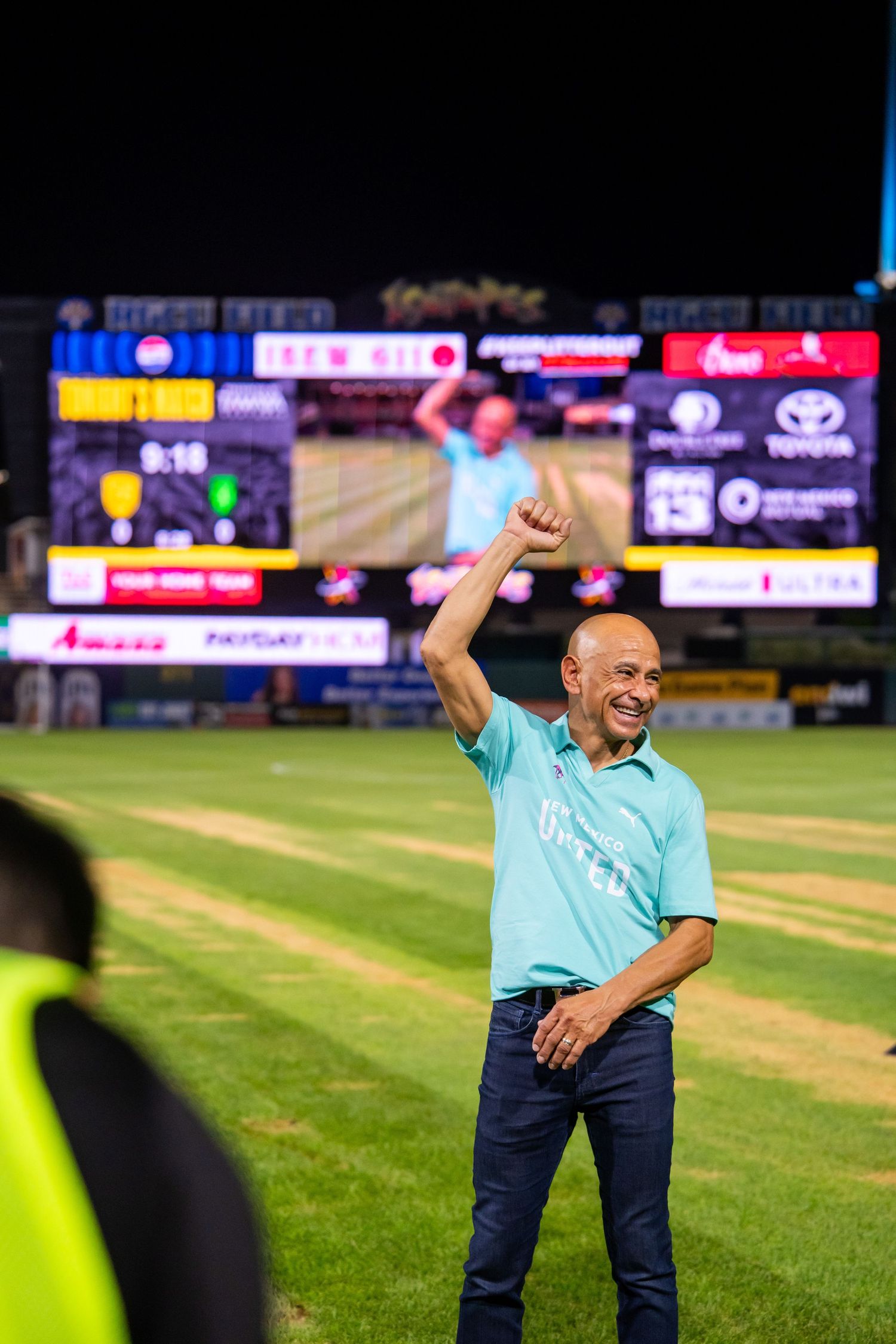 Man in teal shirt celebrates with raised fist on baseball field at night with scoreboard behind him.