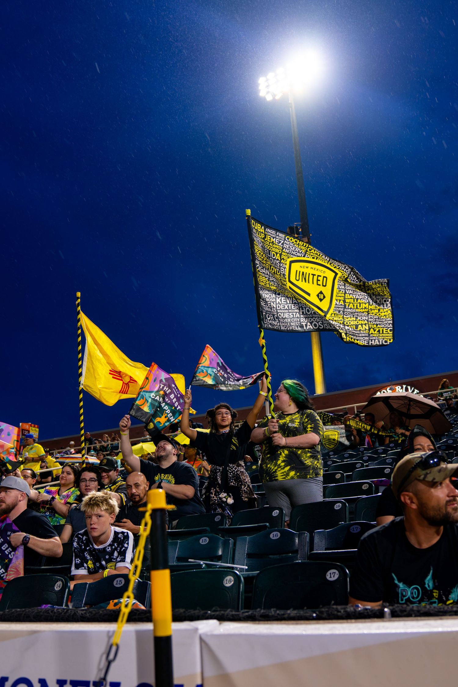 New Mexico United soccer fans wave yellow and black flags in stadium stands under bright floodlights at night.