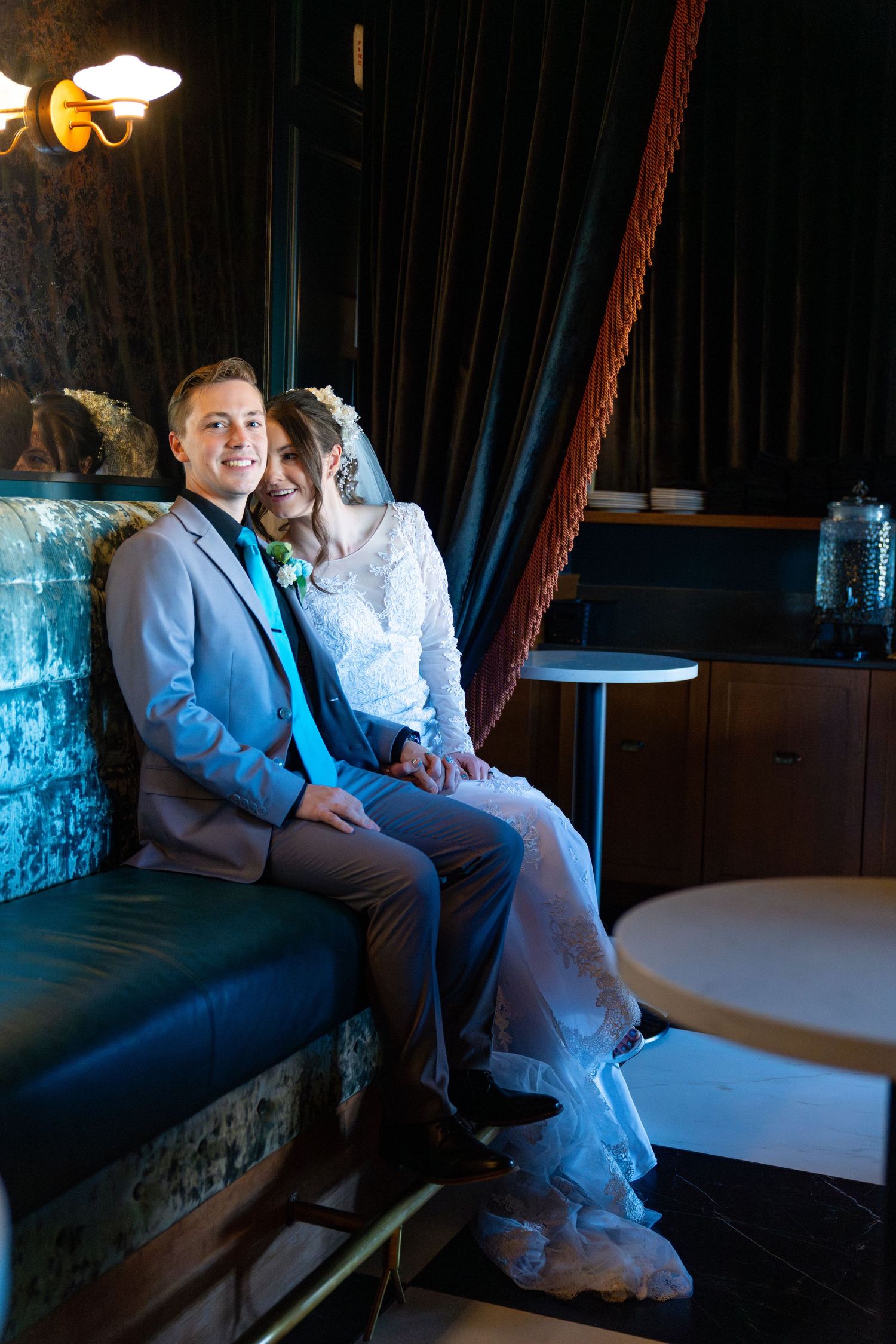 Bride and groom sitting together on a velvet sofa in a dimly lit elegant venue, smiling on their wedding day.