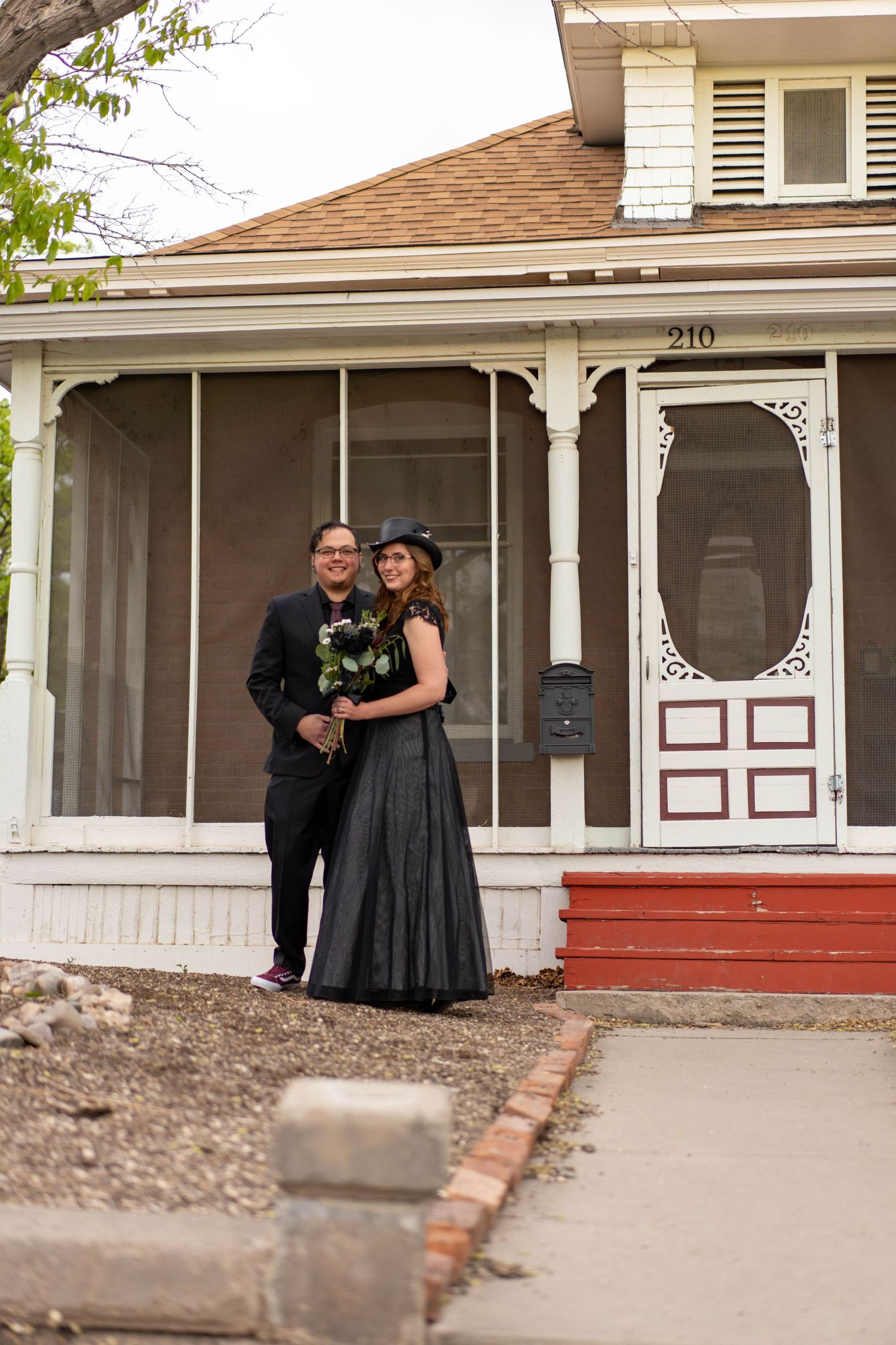 A couple in formal attire share an intimate moment on the porch of a craftsman-style house.