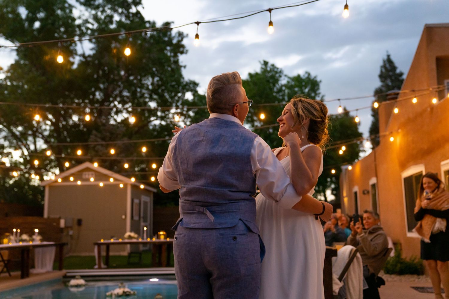 A couple shares their first dance at an outdoor evening reception with string lights glowing overhead in a garden setting.